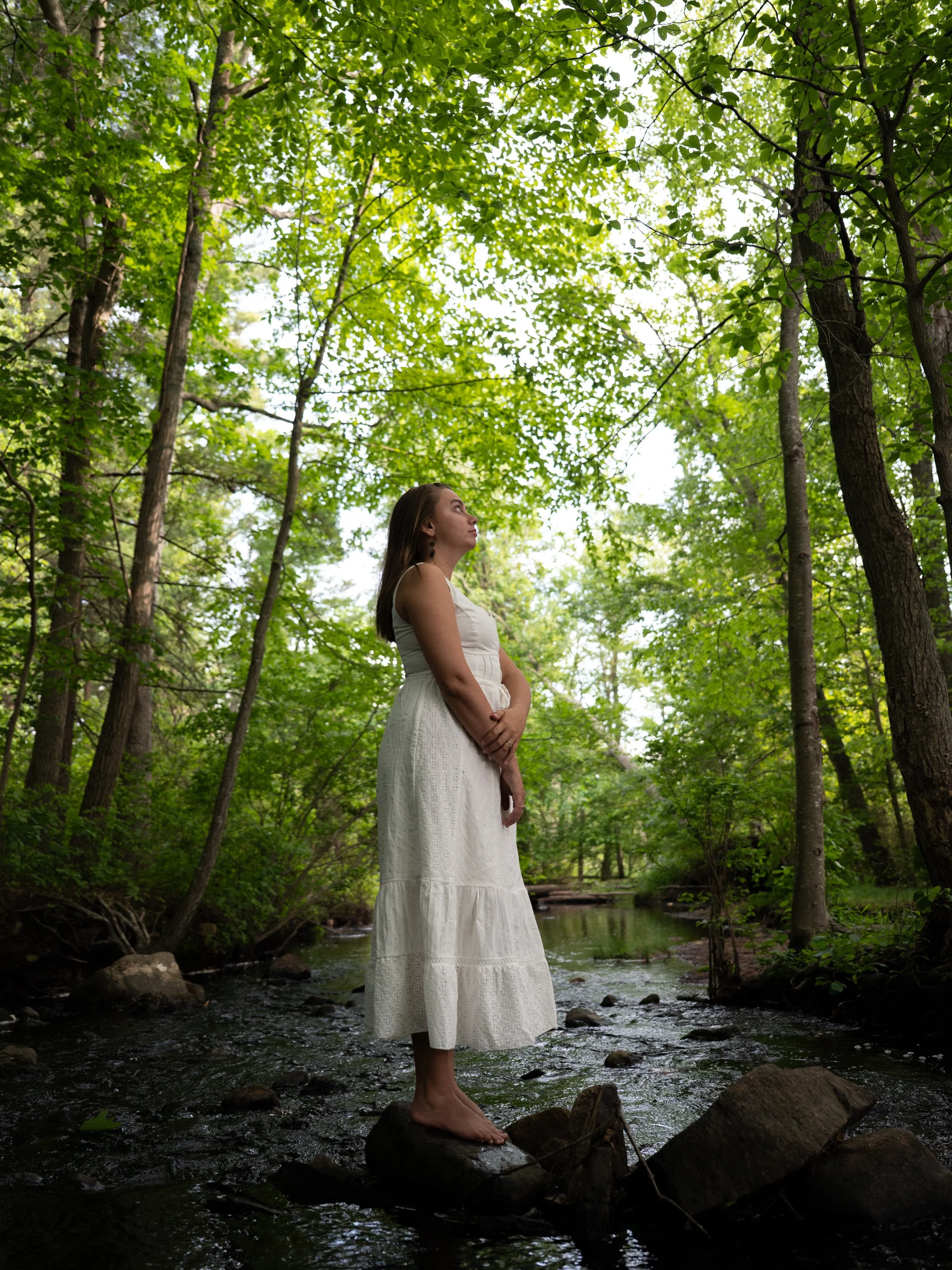 A woman in a white dress standing barefoot on rocks in a flowing creek surrounded by green trees.