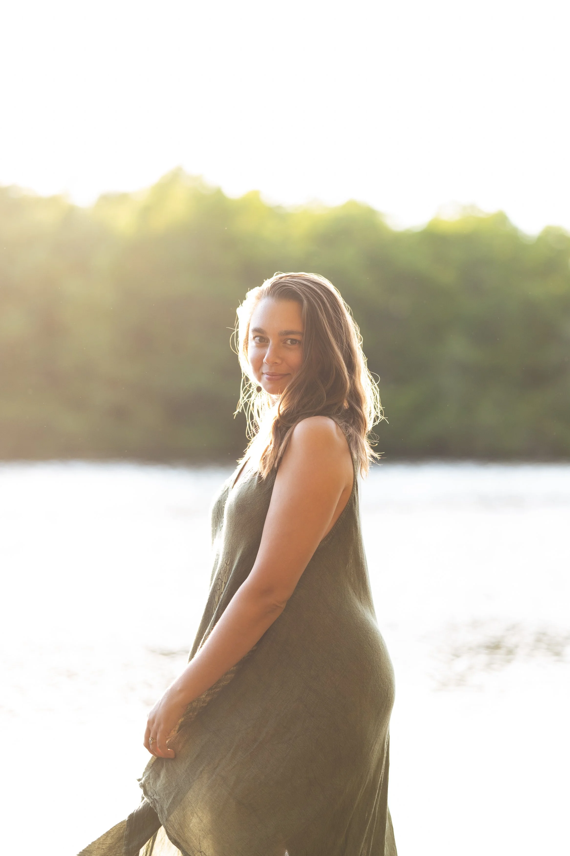 A woman standing outdoors near water, with trees in the background, during bright sunlight. She has long, wavy brown hair and wears a sleeveless, lightweight dress.