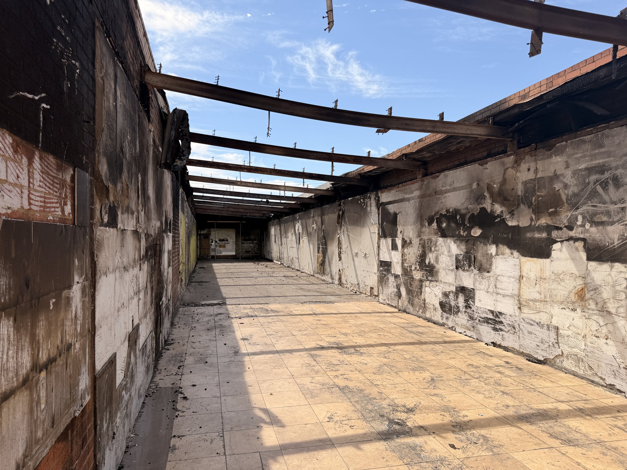 A burnt and partially destroyed rooftop or terrace with charred walls, exposed beams, and debris on the floor, under a clear blue sky.