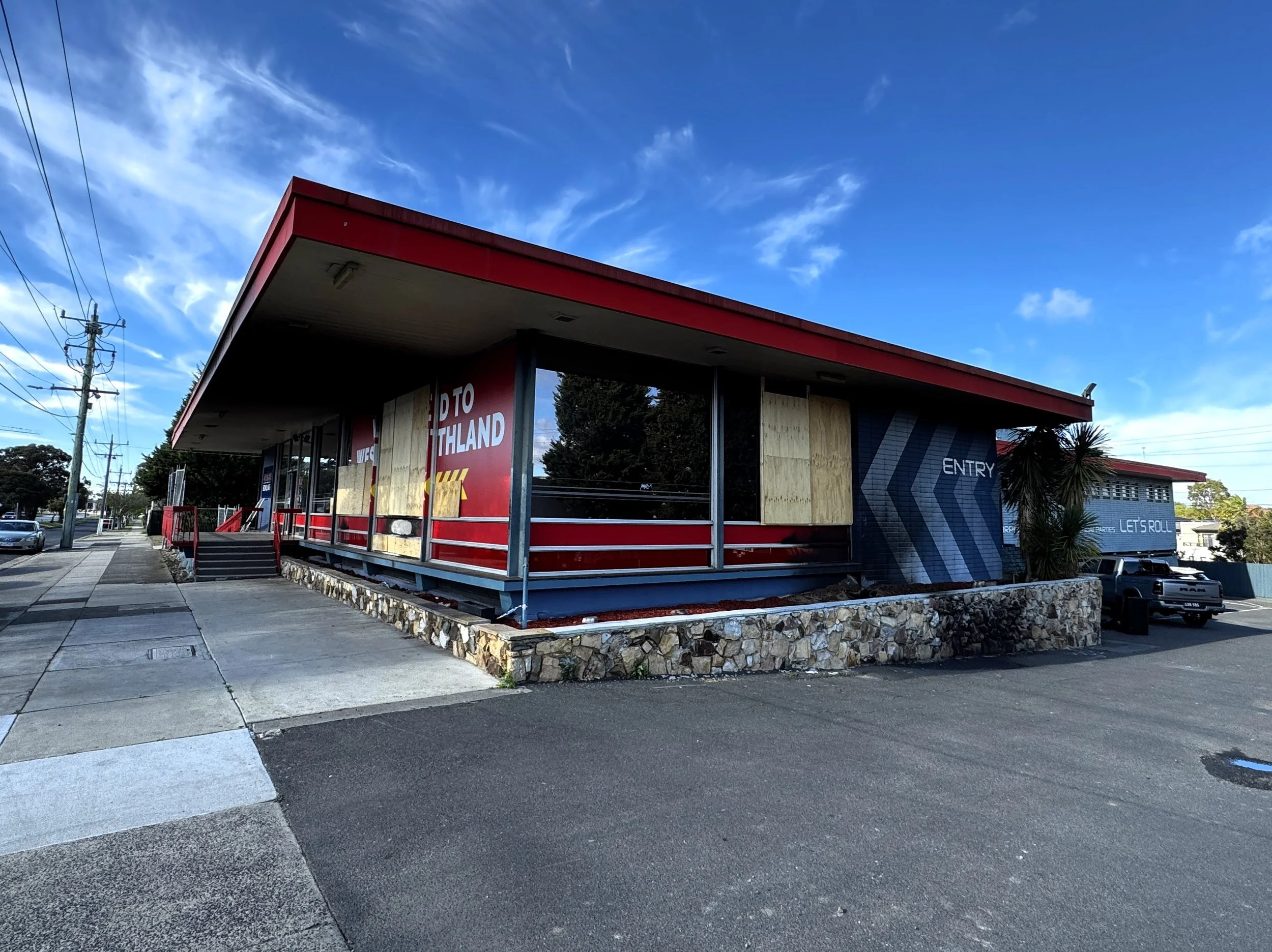 A modern building with a red roof and a mix of glass and wood exterior, displaying signs that read "Welcome to Northland," "Entry," and "Let's Roll." The building is elevated on a stone foundation, with a sidewalk in front and clear sky overhead.