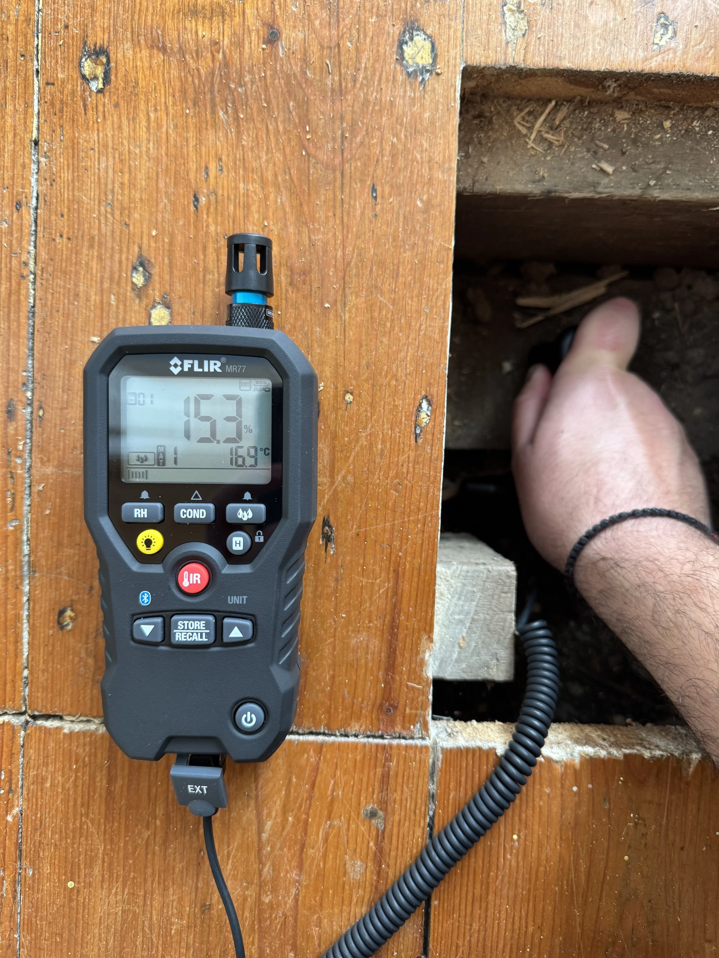 A handheld infrared thermometer displays a temperature of 16.9°C and humidity of 15.3%, placed on a wooden surface near a person using a tool inside a small opening in the wood.