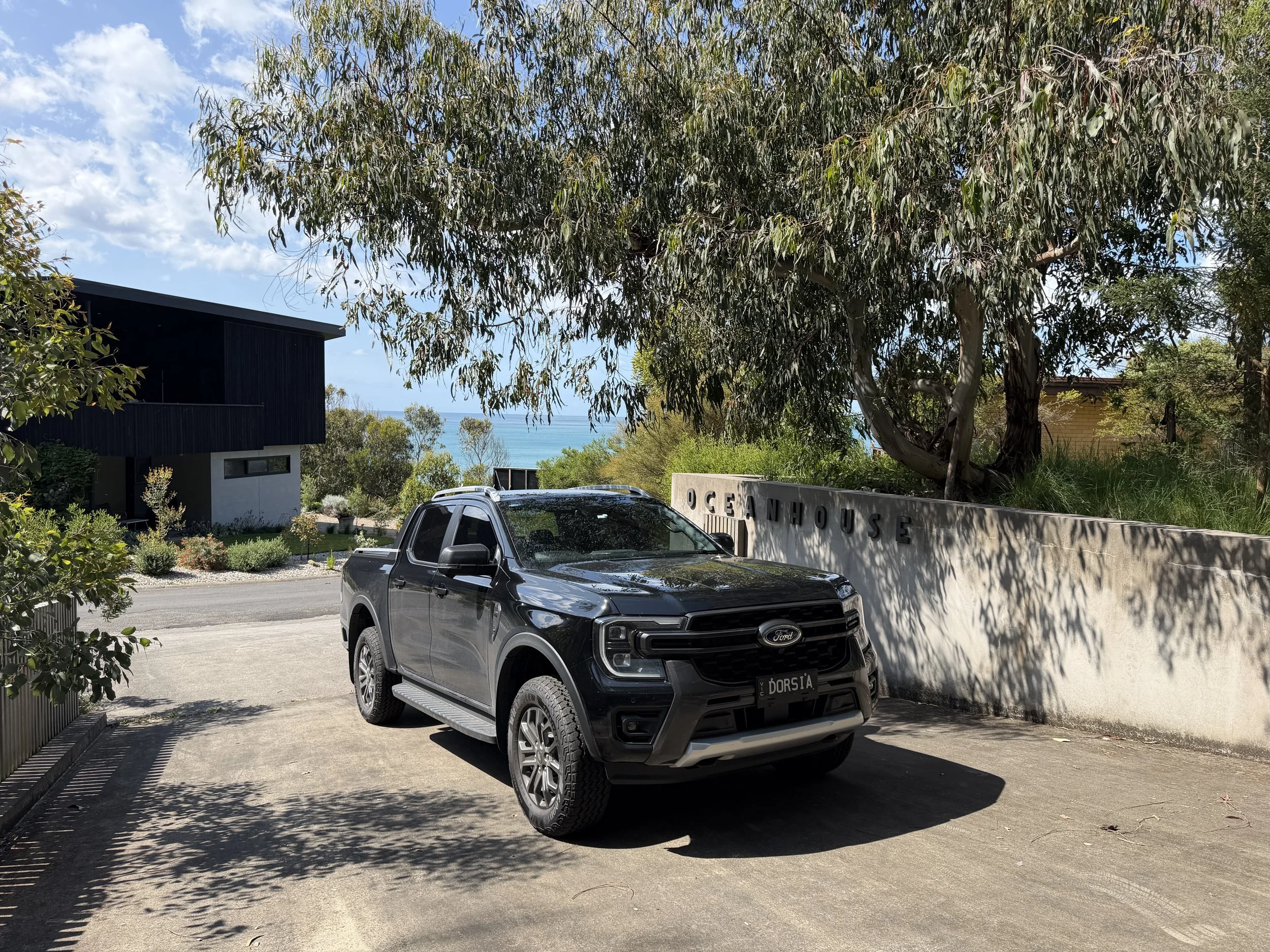 Black Ford pickup truck parked in front of a concrete wall with the words 'OCEAN VIEW HOUSE,' with trees and a view of the ocean and sky in the background.