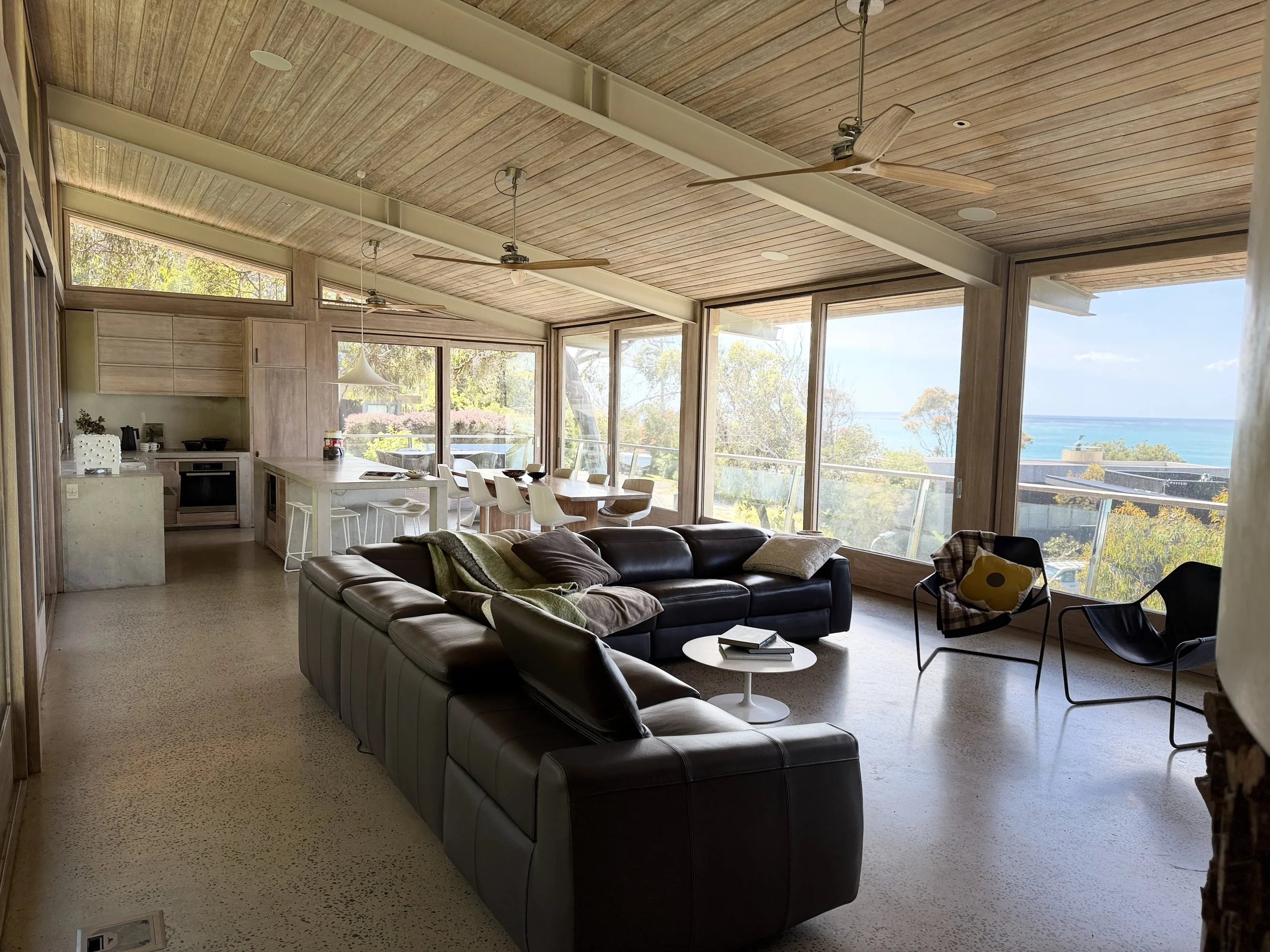 Open-concept living room with large windows showing a view of trees and the ocean, furnished with a black leather sectional sofa, a round white coffee table, and two modern black chairs with cushions. The kitchen area with a large white island and white chairs is visible in the background, with a dining table and chairs near the windows. The ceiling features wood paneling and ceiling fans.