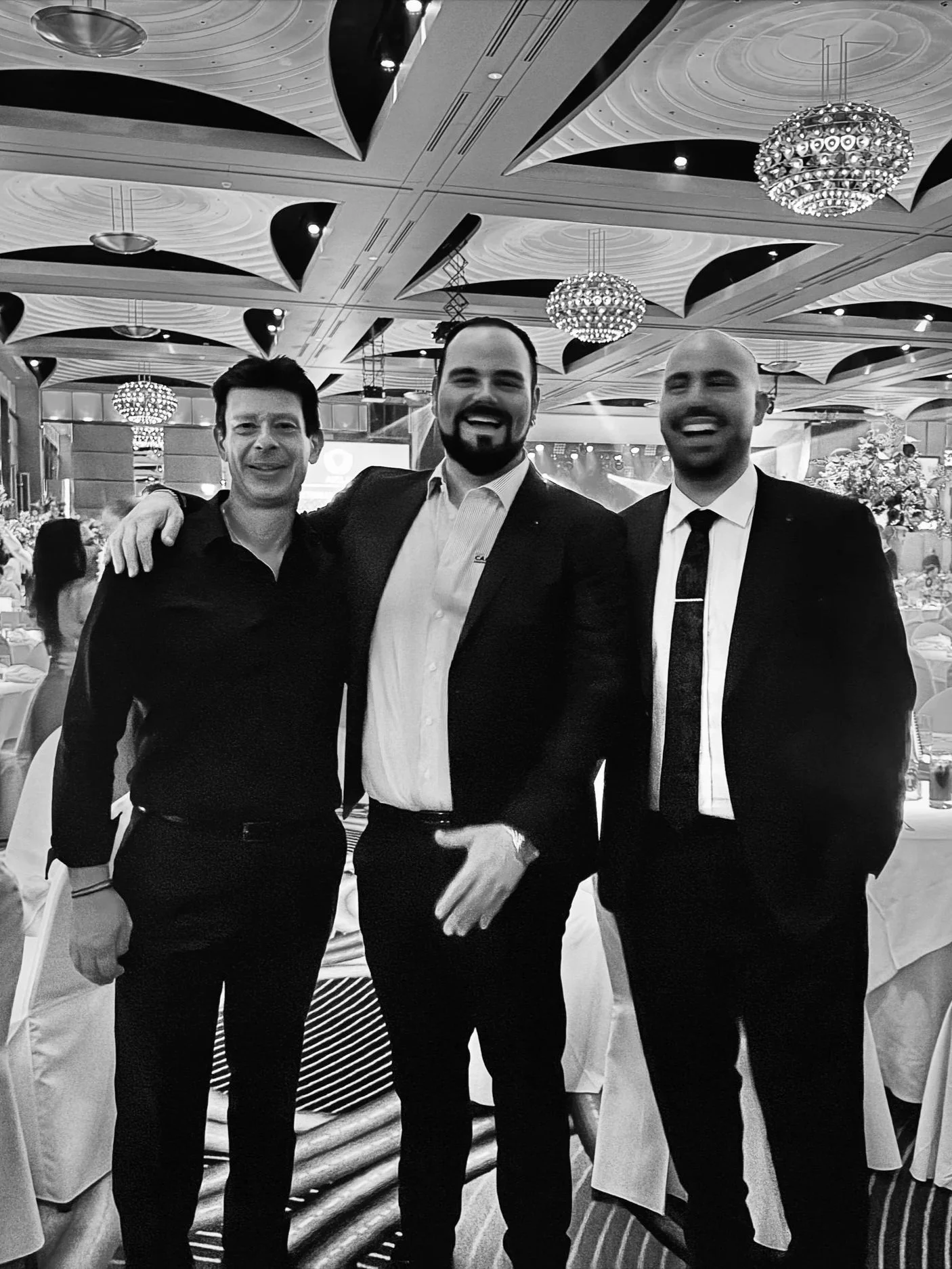 Three men in formal attire smiling and posing together at an indoor event. The background shows chandeliers, decorated tables, and a large banquet hall.