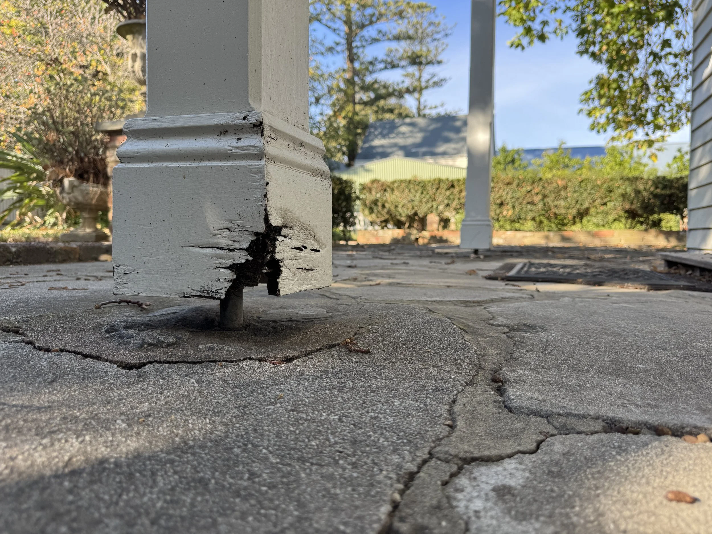 Close-up of a cracked and peeling white wooden porch post on a stone porch, with some plants and bushes in the background.