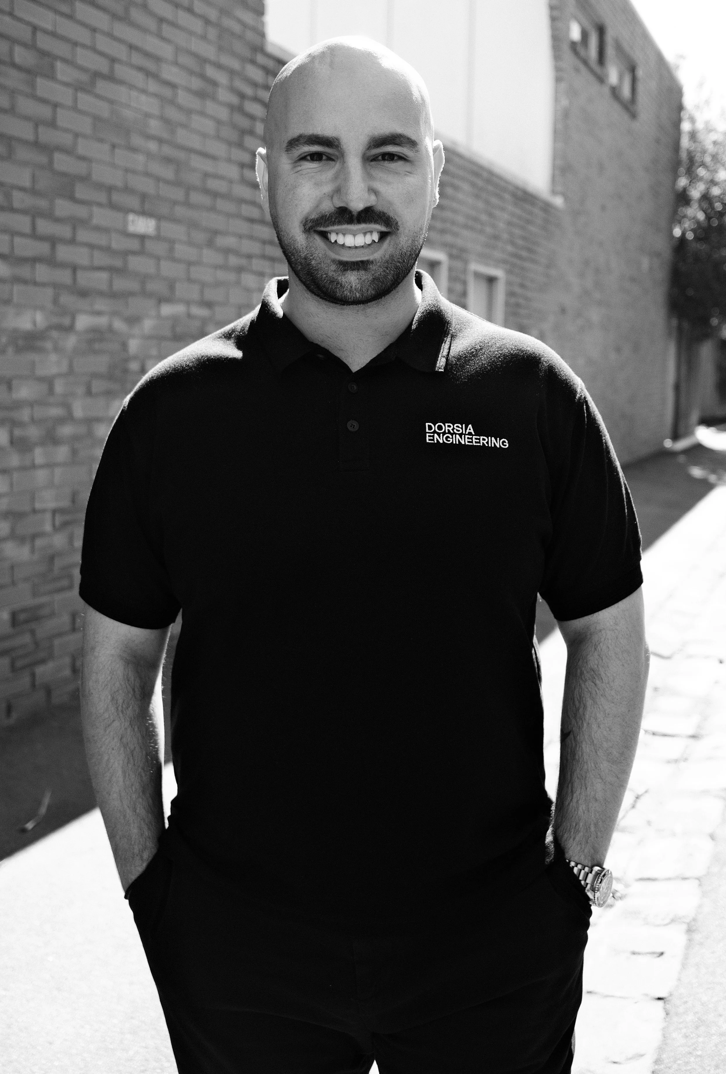A smiling man with a beard and shaved head, wearing a black polo shirt that has "DORSIA ENGINEERING" embroidered on it, standing outdoors next to a brick building with his hands in his pockets.