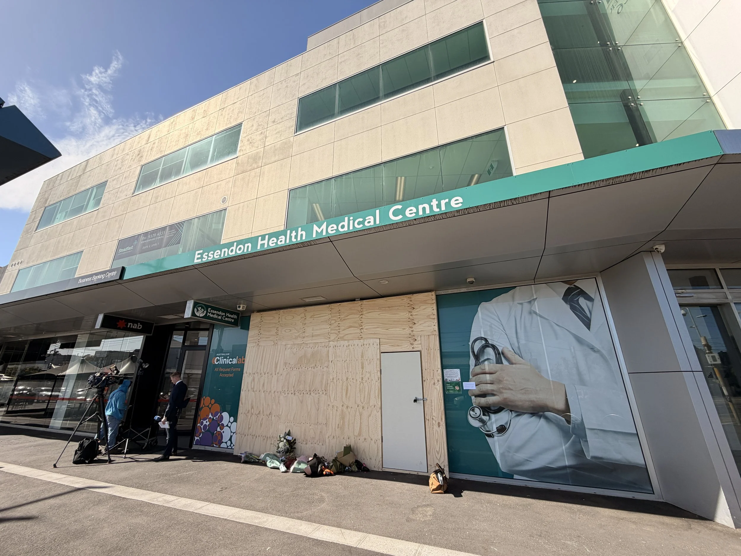 The entrance of Essendon Health Medical Centre with a corner poster of a doctor, and some flowers and items placed on the ground in front, possibly a memorial.