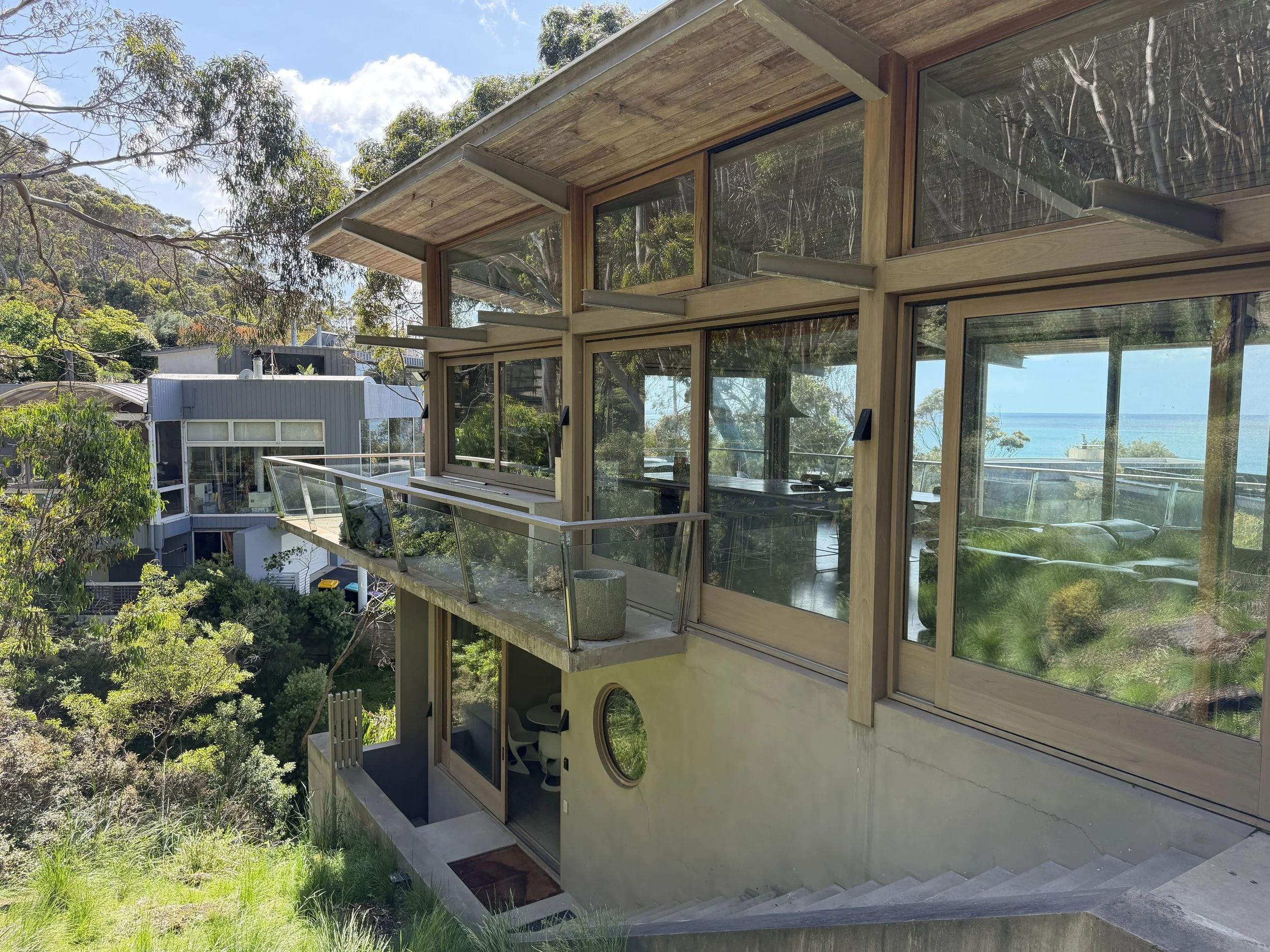 Modern house with large glass windows and balcony, surrounded by trees, with ocean view in the background.