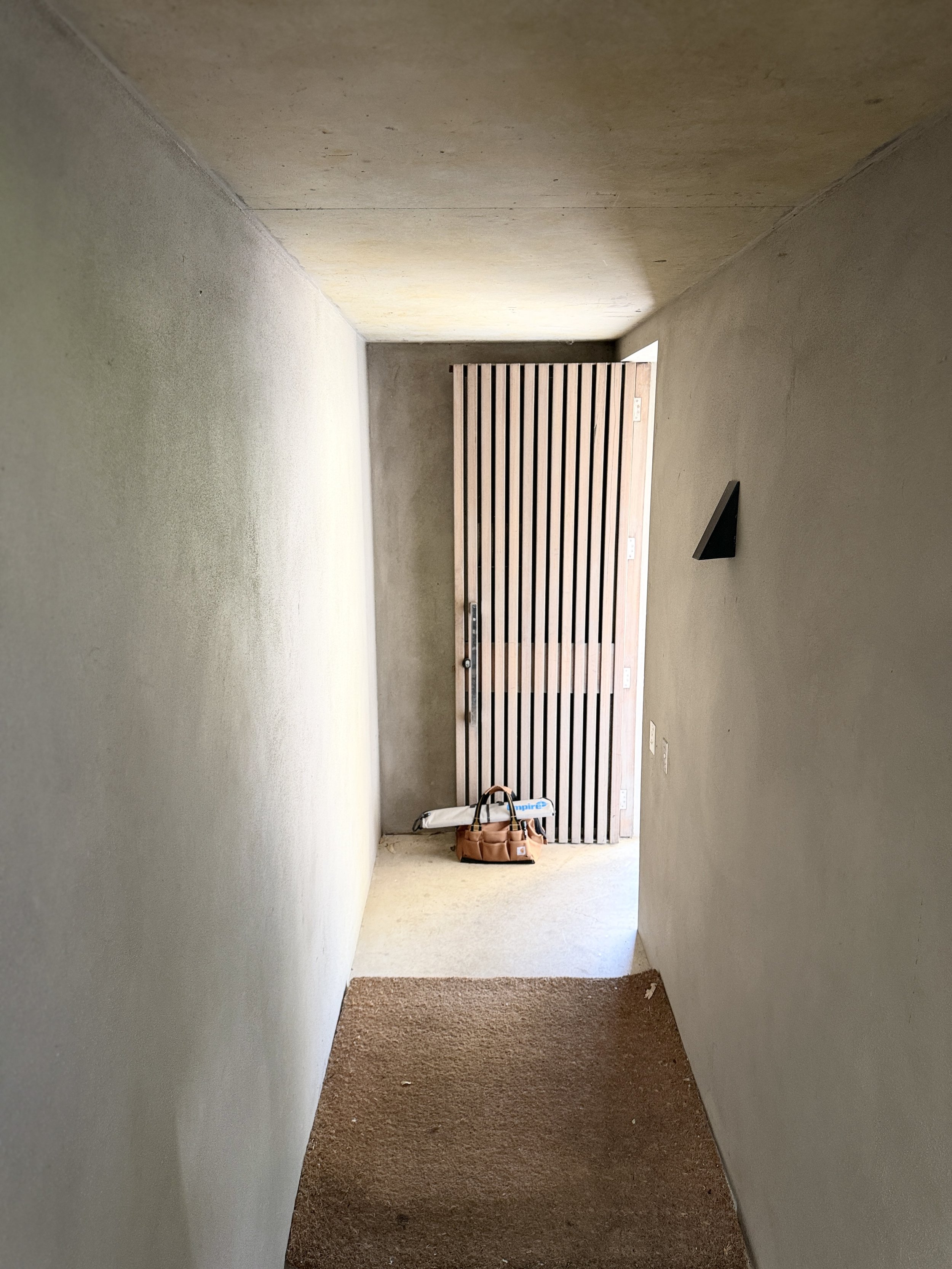 Entrance of a narrow hallway with a brown carpet at the beginning leading to a concrete floor and a wooden folding door, with tools and a bag on the floor near the door.
