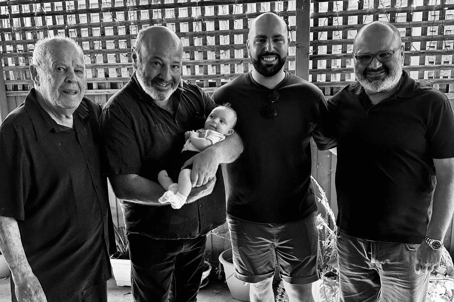 Five men standing together in a line, with one holding a baby, in a wooden structure with lattice walls.