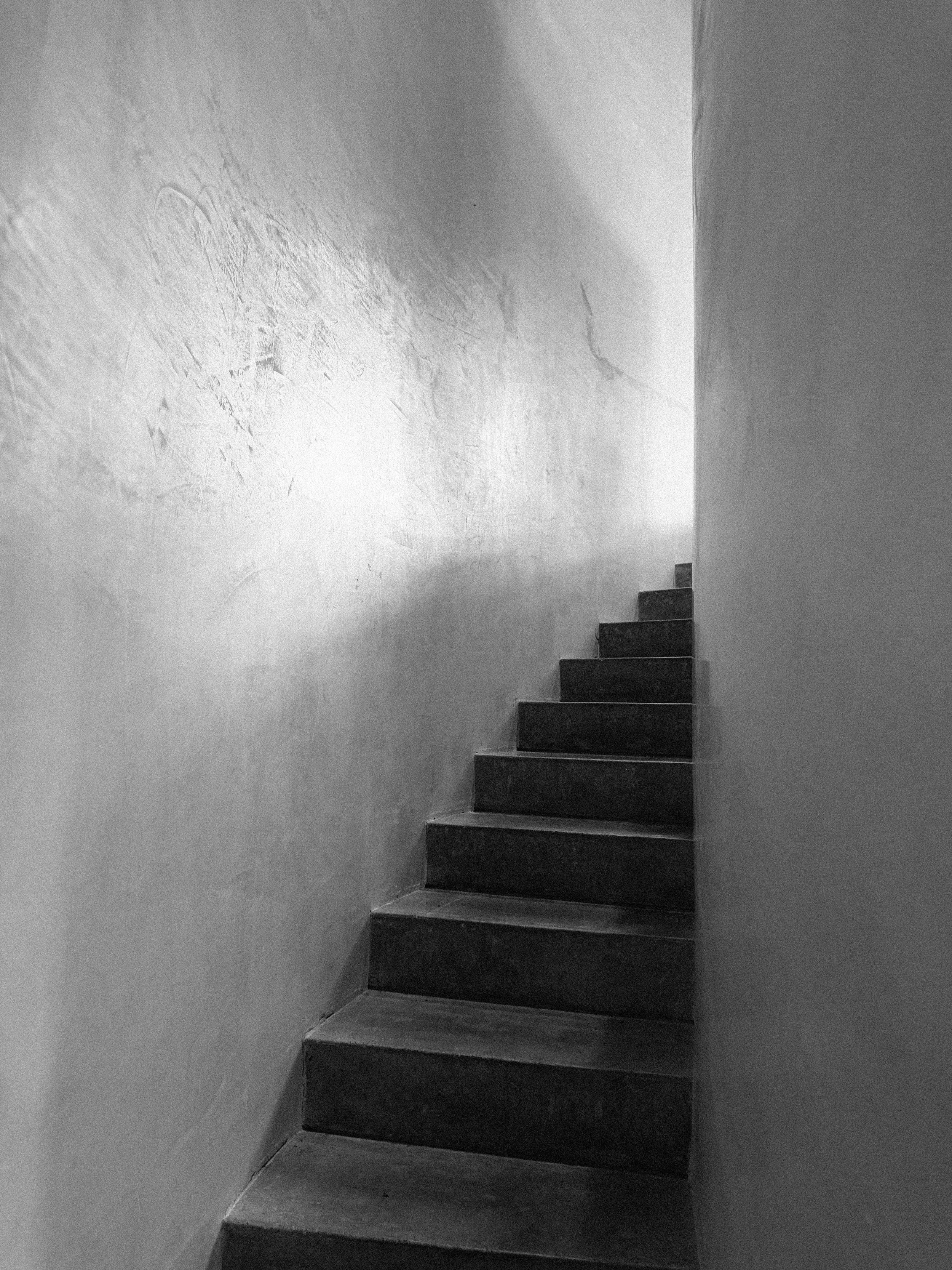 Black and white photo of a narrow staircase with dark steps, enclosed by plain walls, leading upward into an lighted area.