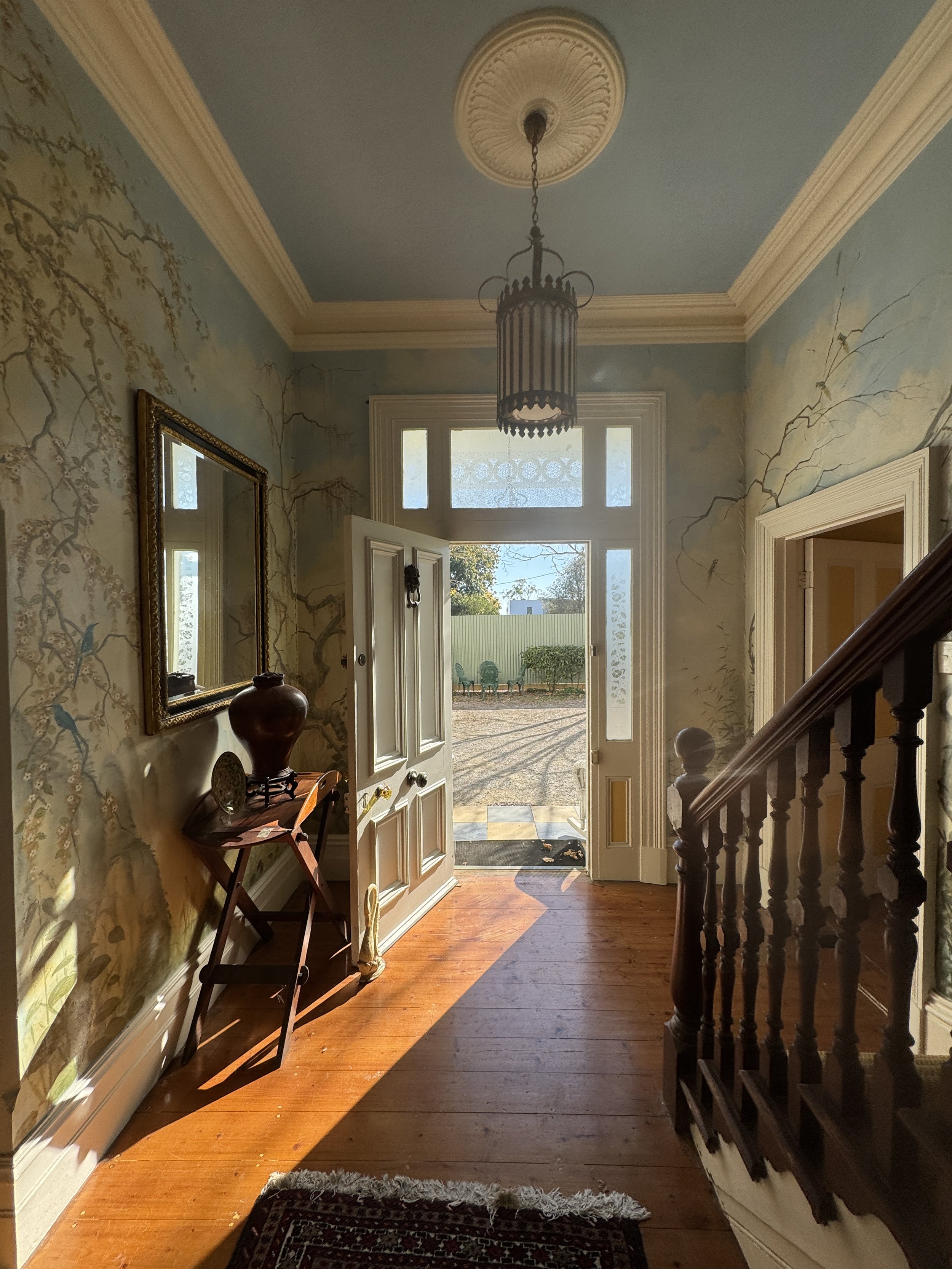 Entryway with wooden floor, an open front door, a mirror on the wall, a small table with a vase, stairs on the right, and sunlight streaming inside.