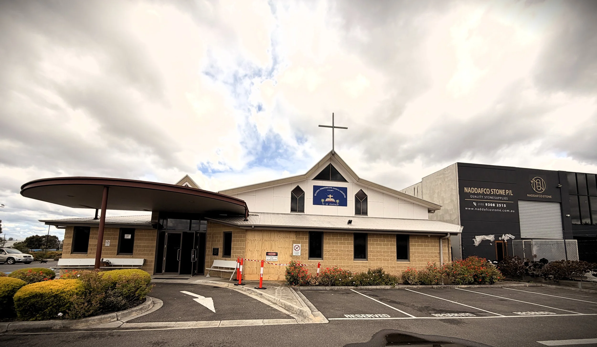 A church-style building with a cross on top, surrounded by a parking lot with reserved parking spaces, bushes, and a cloudy sky above.