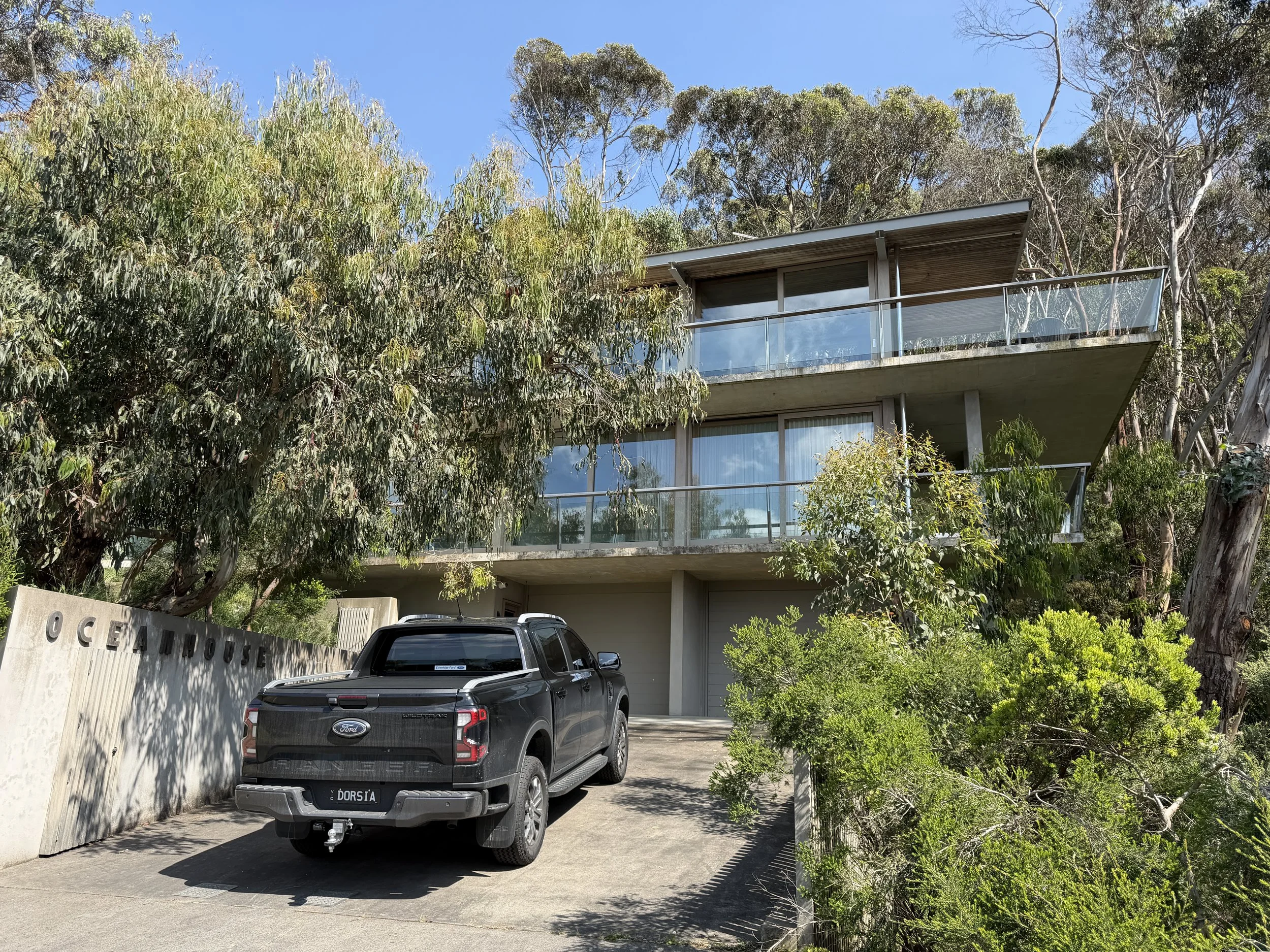 Modern multi-story house with glass balconies, surrounded by trees and greenery, with a black Ford truck parked in the driveway, and a concrete wall on the left side with the word 'Oceanview'.