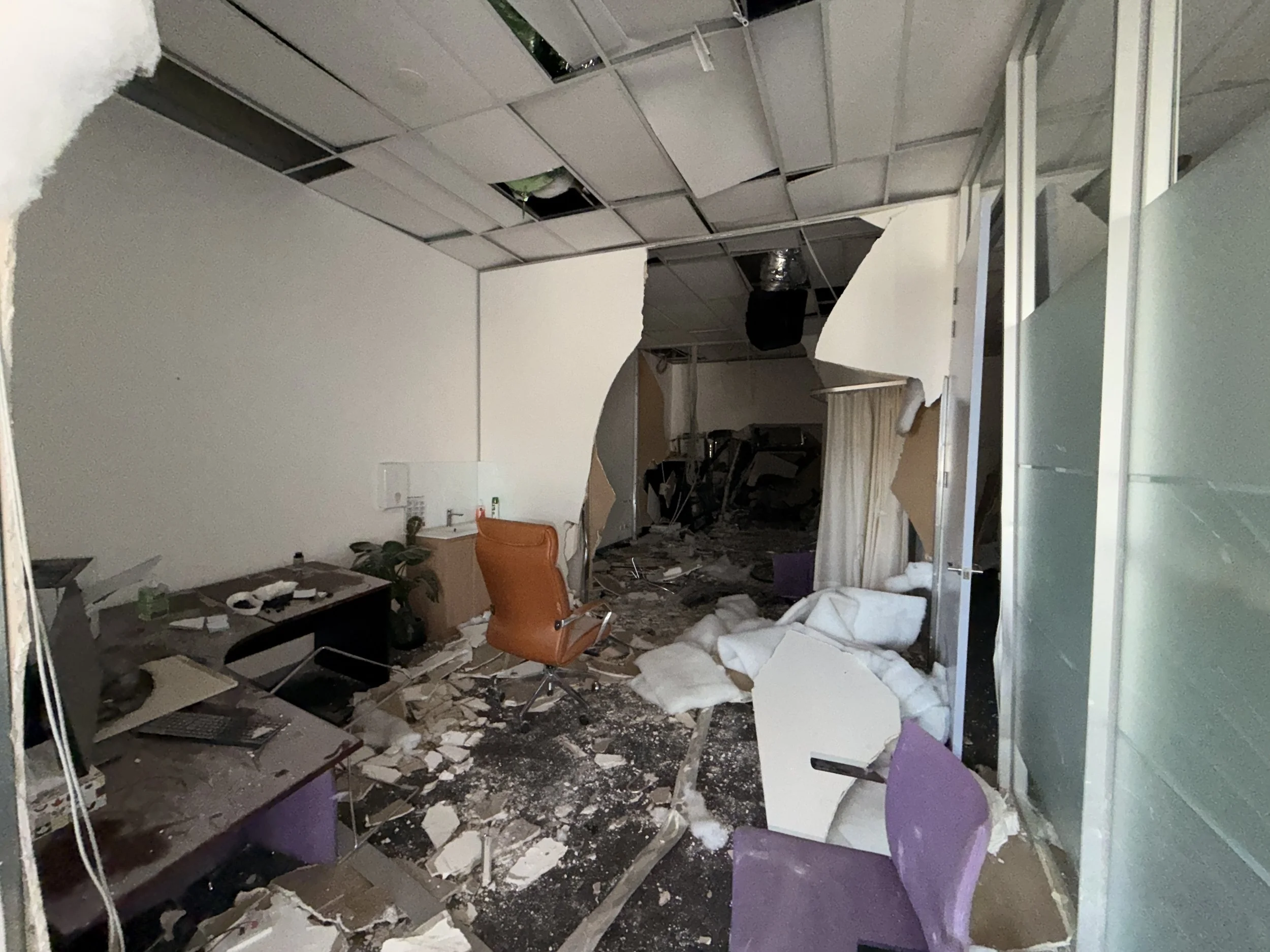 Office room with collapsed ceiling tiles, debris on the floor, broken glass, and damaged furniture including a brown office chair and purple chairs, with a desk and workplace equipment.