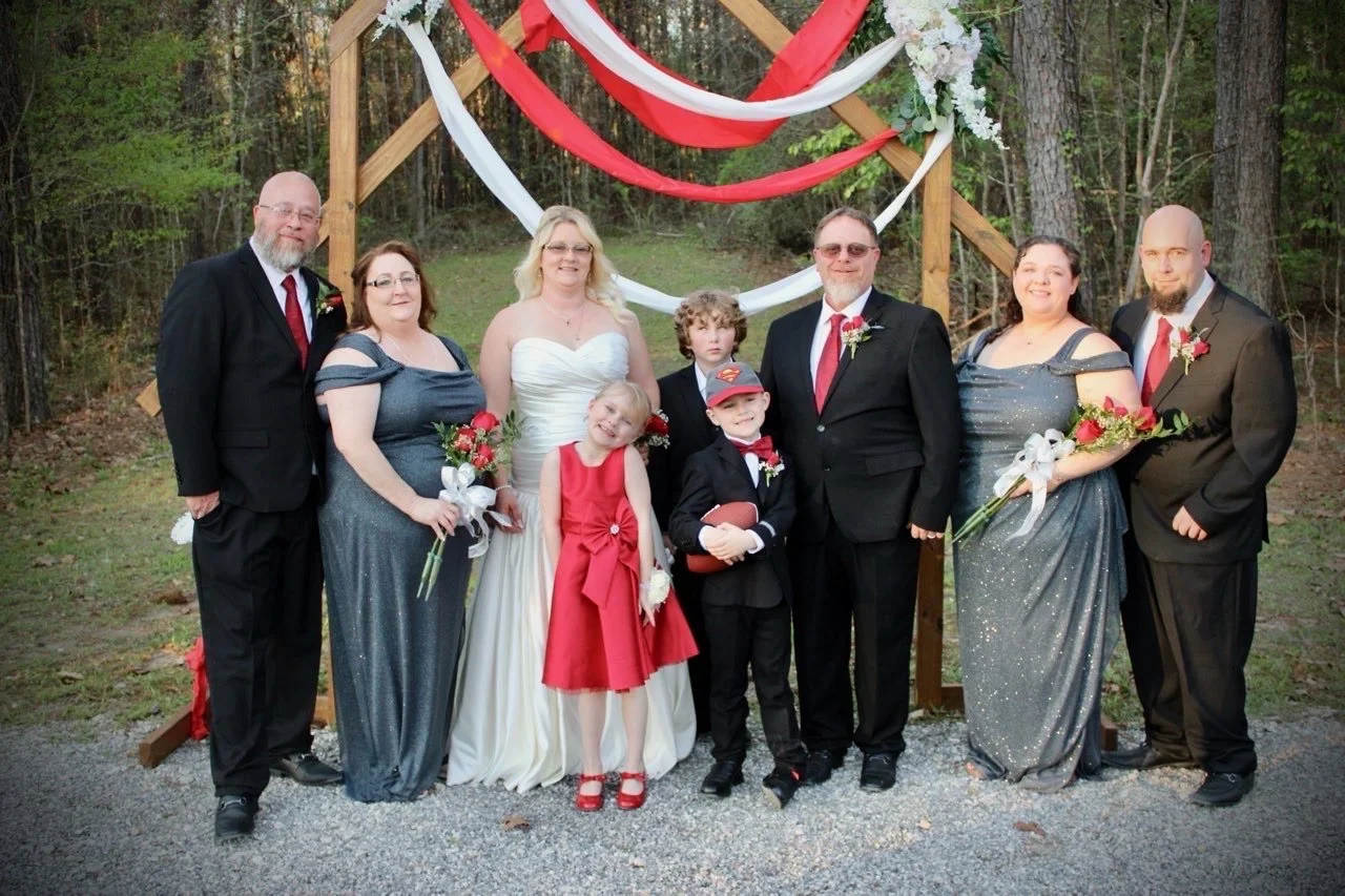 Group of people at a wedding outdoors in front of a wooden arch decorated with red and white fabric and flowers, surrounded by trees.