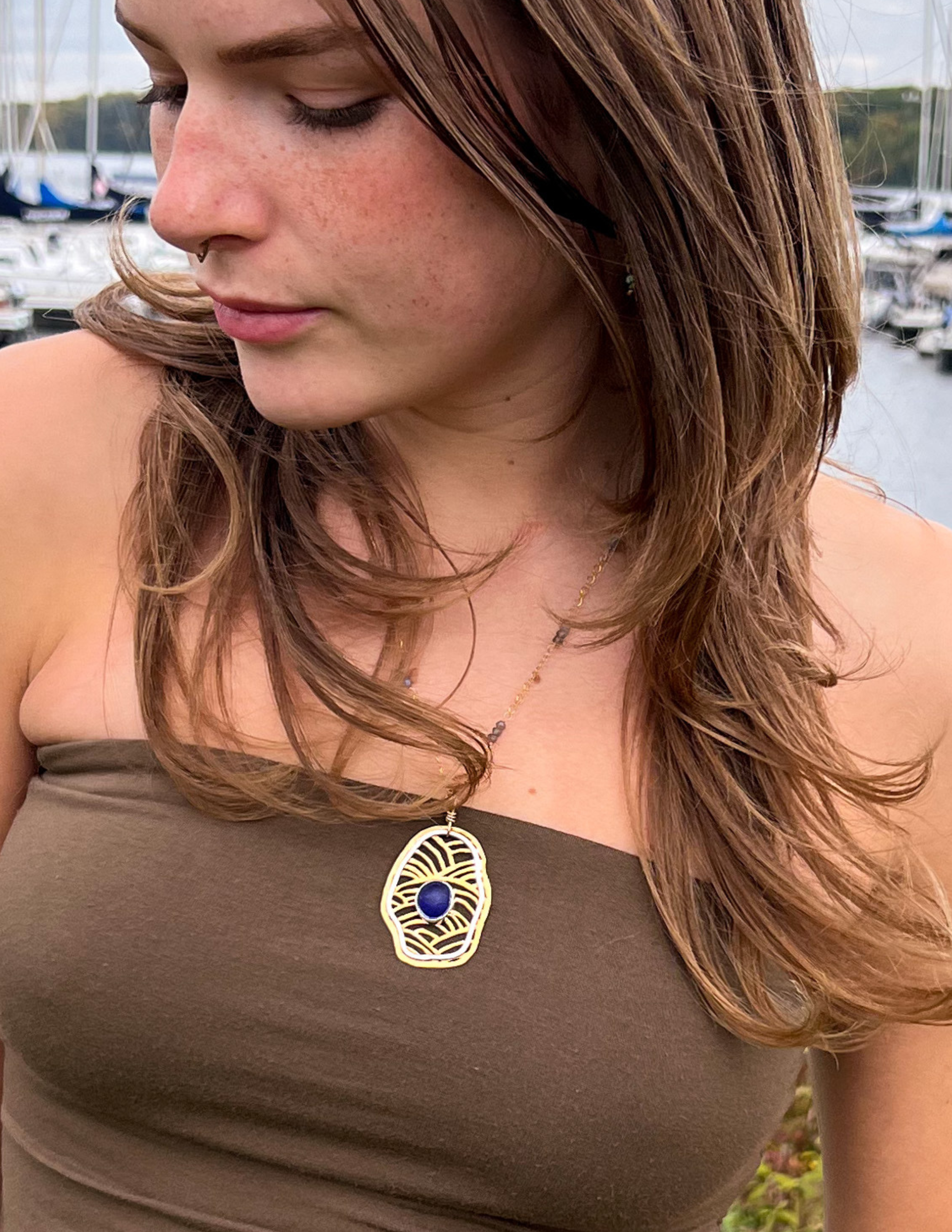 Young woman looking down wearing a n organically shaped saw pierced brass pendant necklace that has silver accents and a cobalt blue sea glass cabochon.