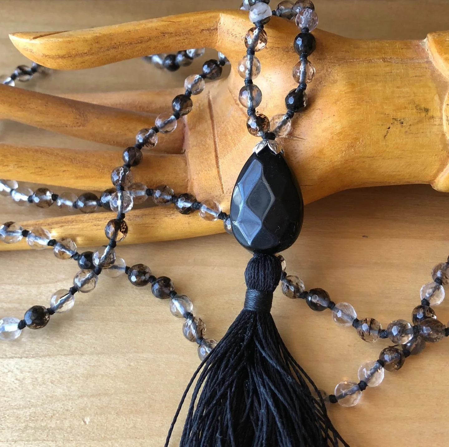 A black beaded necklace with a large teardrop-shaped pendant, attached to a black tassel, resting on a wooden sculpture.