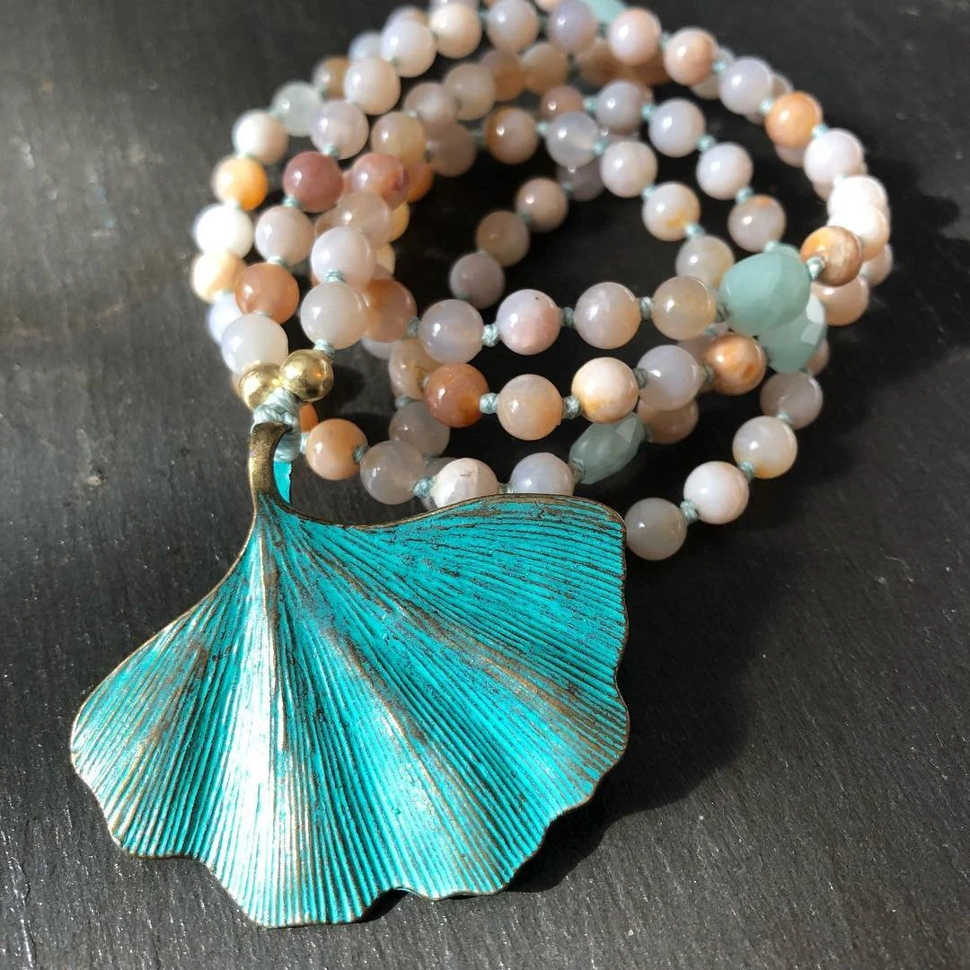 A beaded necklace with round pink and white pearls, some green beads, and a large, teal, fan-shaped metal pendant with gold accents, resting on a dark surface.