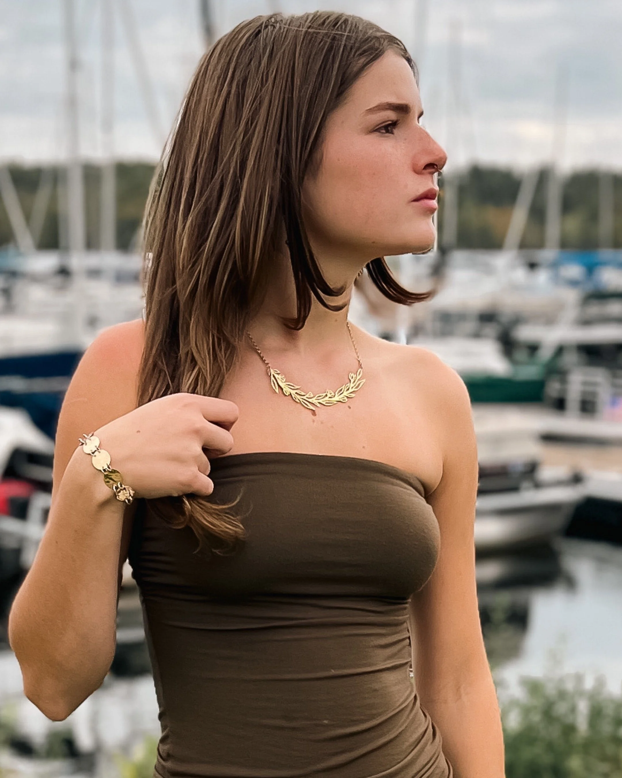 A woman with brown hair wearing a strapless dark brown top, gold jewelry including a necklace and bracelet, standing near a marina with boats and water in the background.