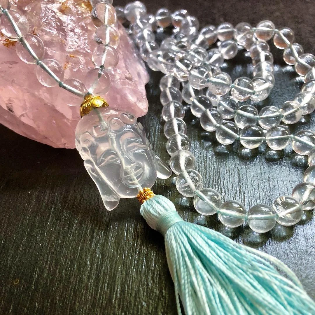 A beaded mala with transparent beads, a carved clear Buddha pendant, a tassel, and a pink crystal rock on a black surface.