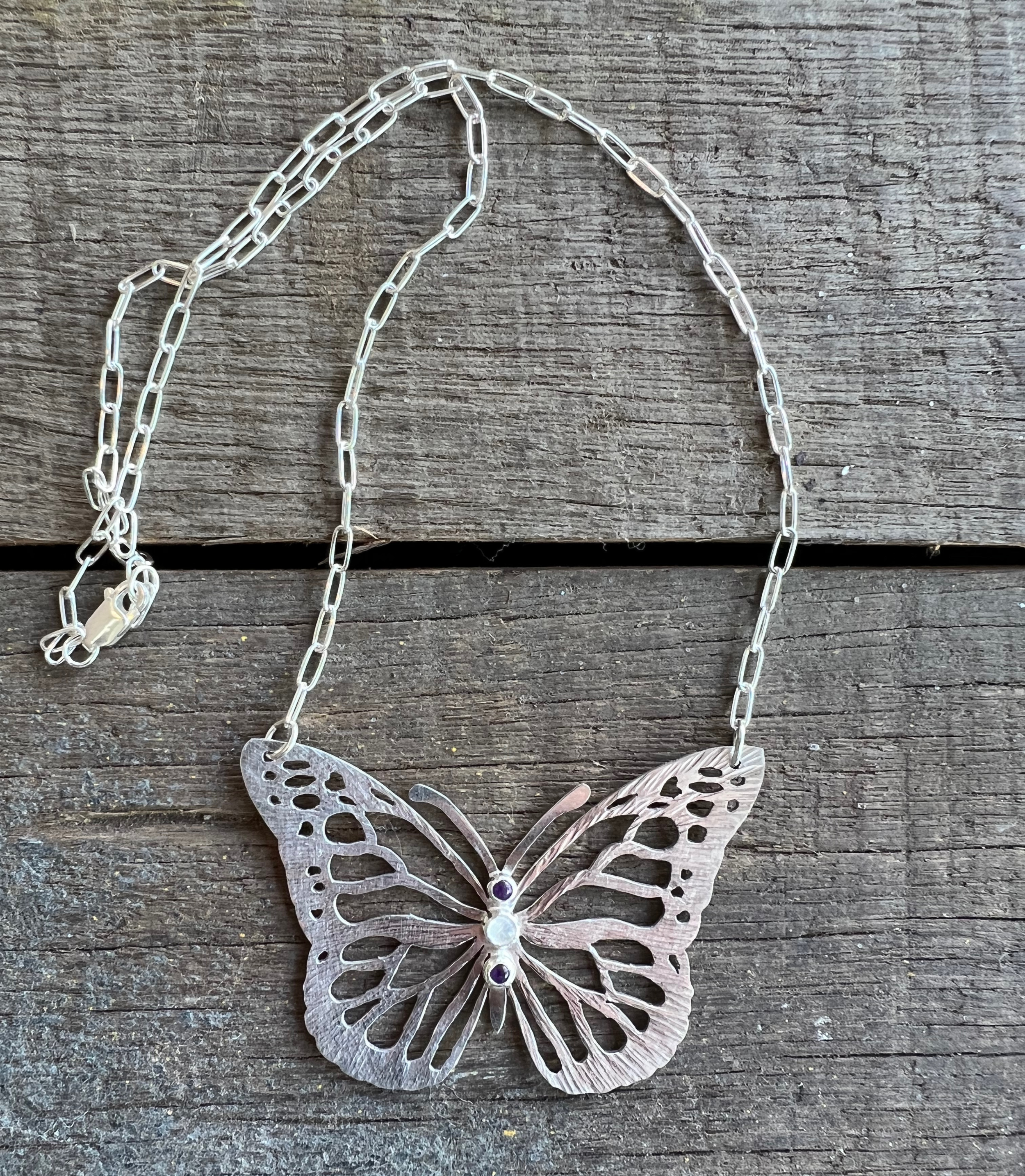 A silver butterfly necklace with intricate cut-out details, laid on a rustic wooden surface.