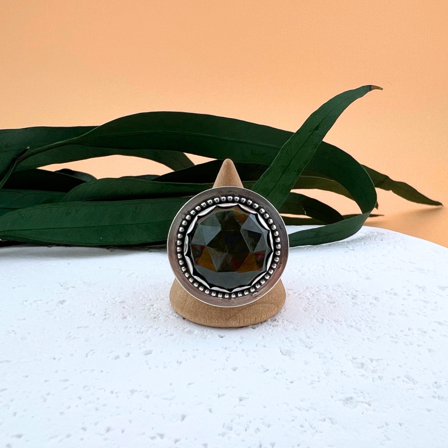 Front view of a sterling silver velvet obsidian ring displayed on a wooden ring stand atop a white stone, highlighting the stone’s faceted surface and deep tone.