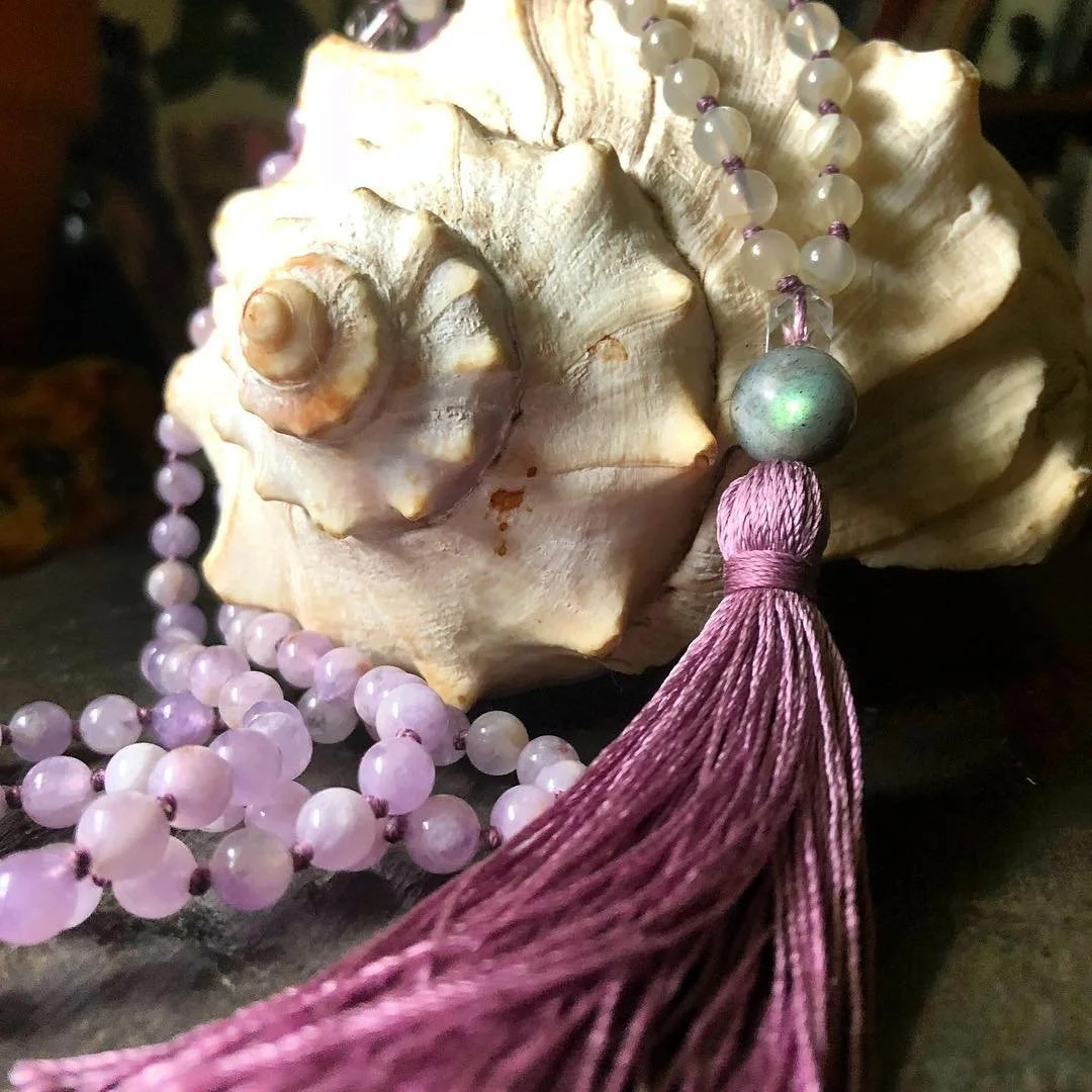 A strand of pink and white beaded necklace with a large, shiny, metallic green bead and purple tassel, draped over a seashell with a spiral shape and rough texture.