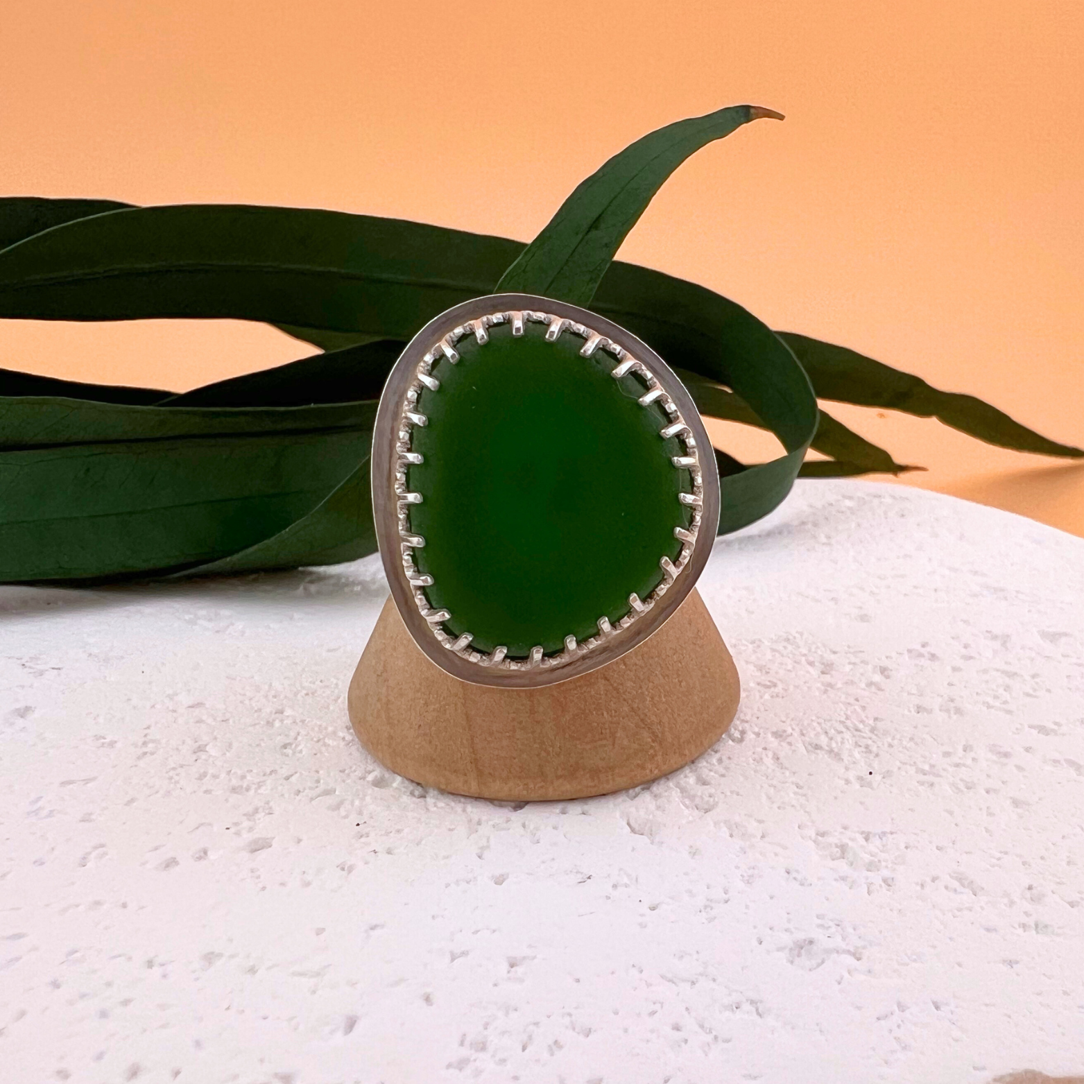 Bright green sea glass ring set in sterling silver, displayed on a white stone surface with green foliage against a warm background.