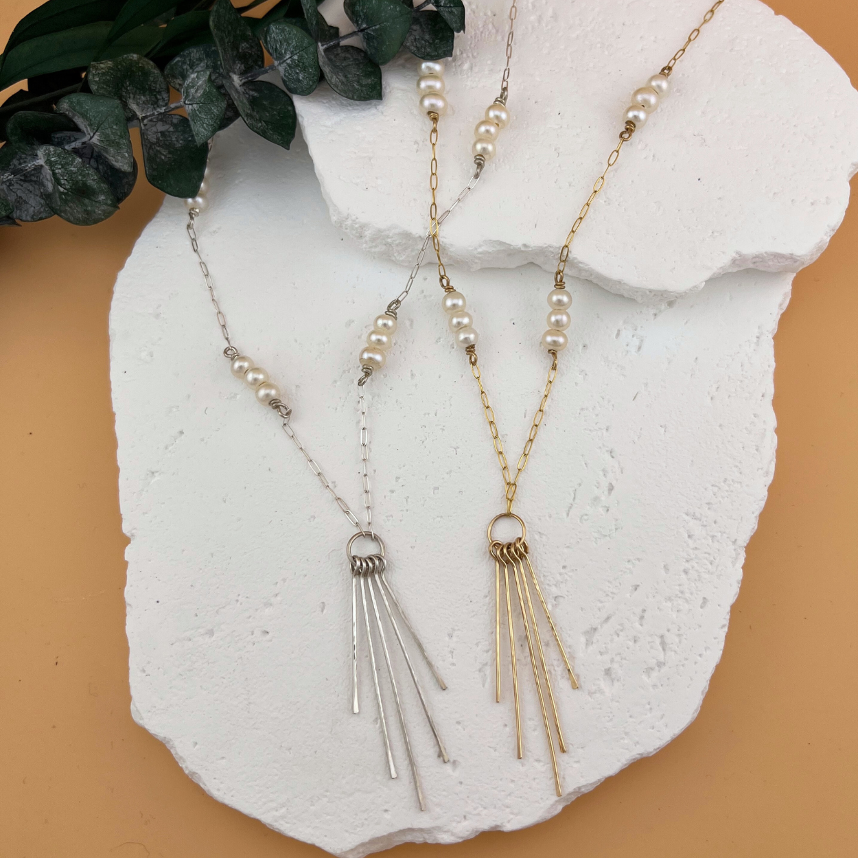 Sterling silver and gold fill Lumen Fringe Necklaces with AAA freshwater pearls displayed on a white stone backdrop, featuring delicate cascading fringe pendants..