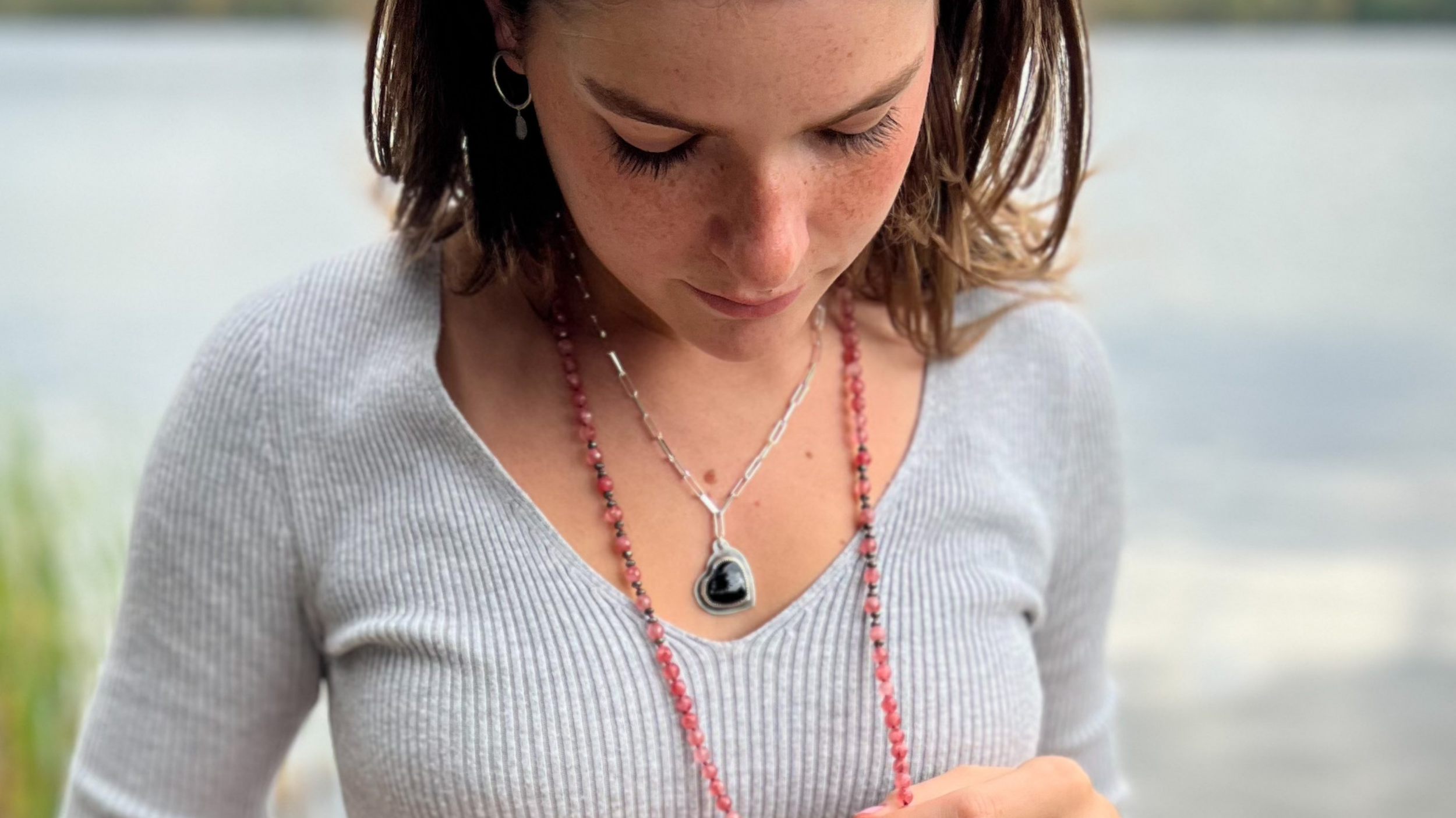 A young woman outside wearing an onyx heart necklace looking down at a strawberry quartz mala that she is holding in her hands.