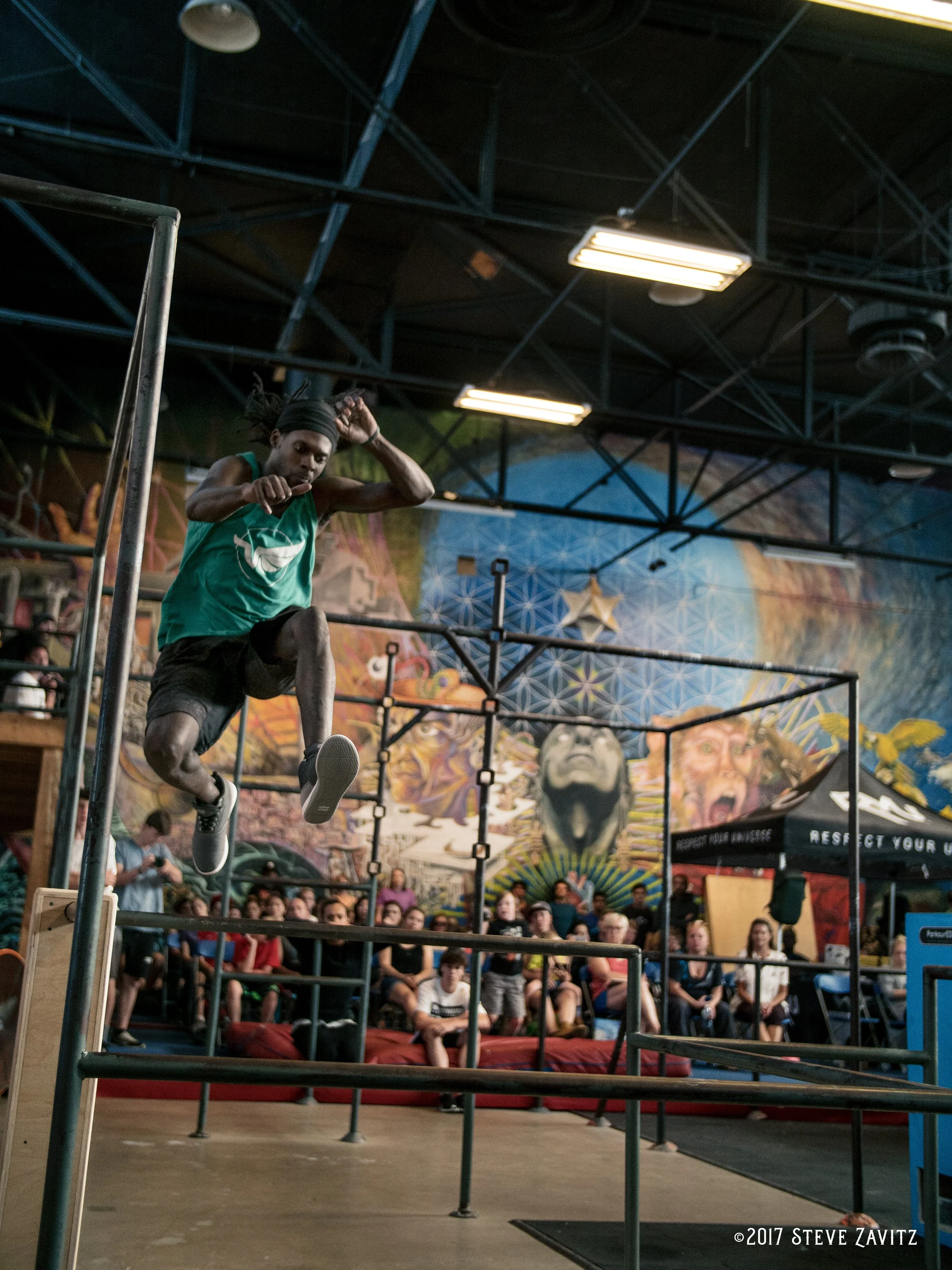 An athlete in a green sleeveless shirt and black shorts is climbing over a hurdle during an obstacle course event inside a gym with a large, colorful mural on the wall behind. The audience is seated in the background.
