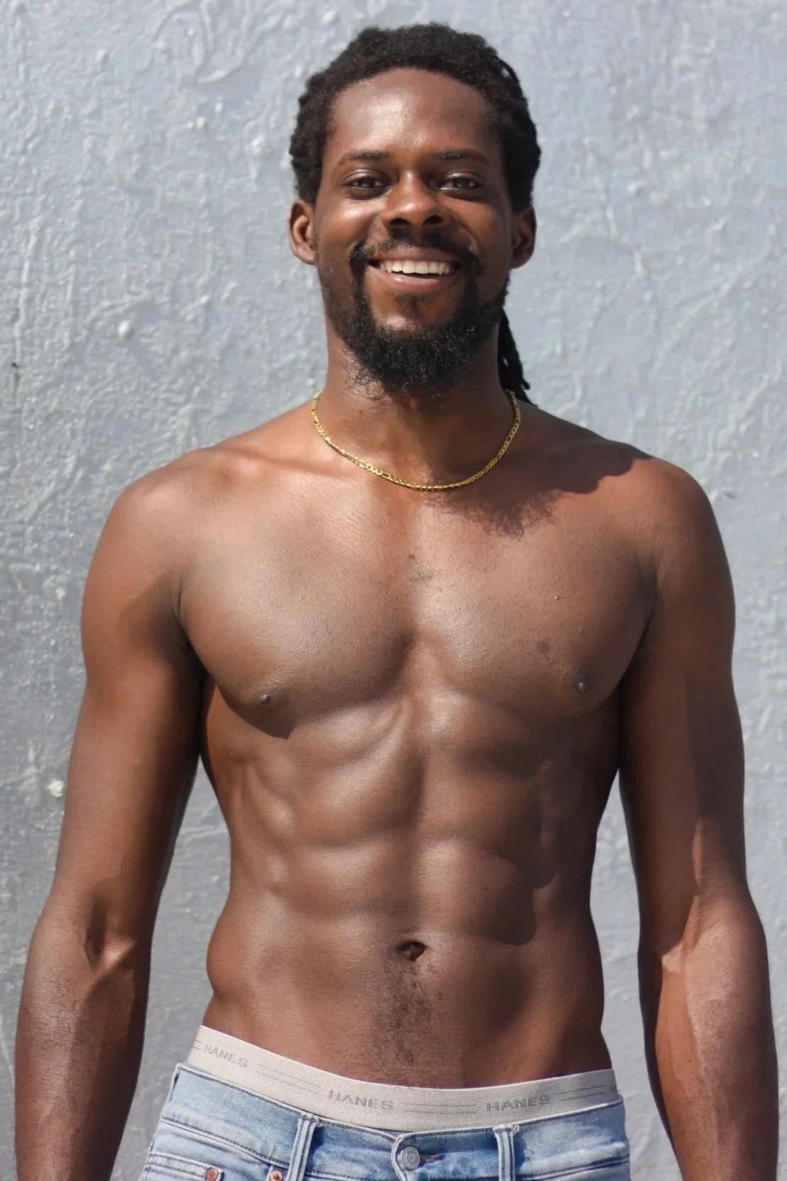 A shirtless man with a muscular build, smiling, standing against a textured gray wall. He has dark skin, a beard, dark curly hair, and is wearing a gold chain necklace and jeans.