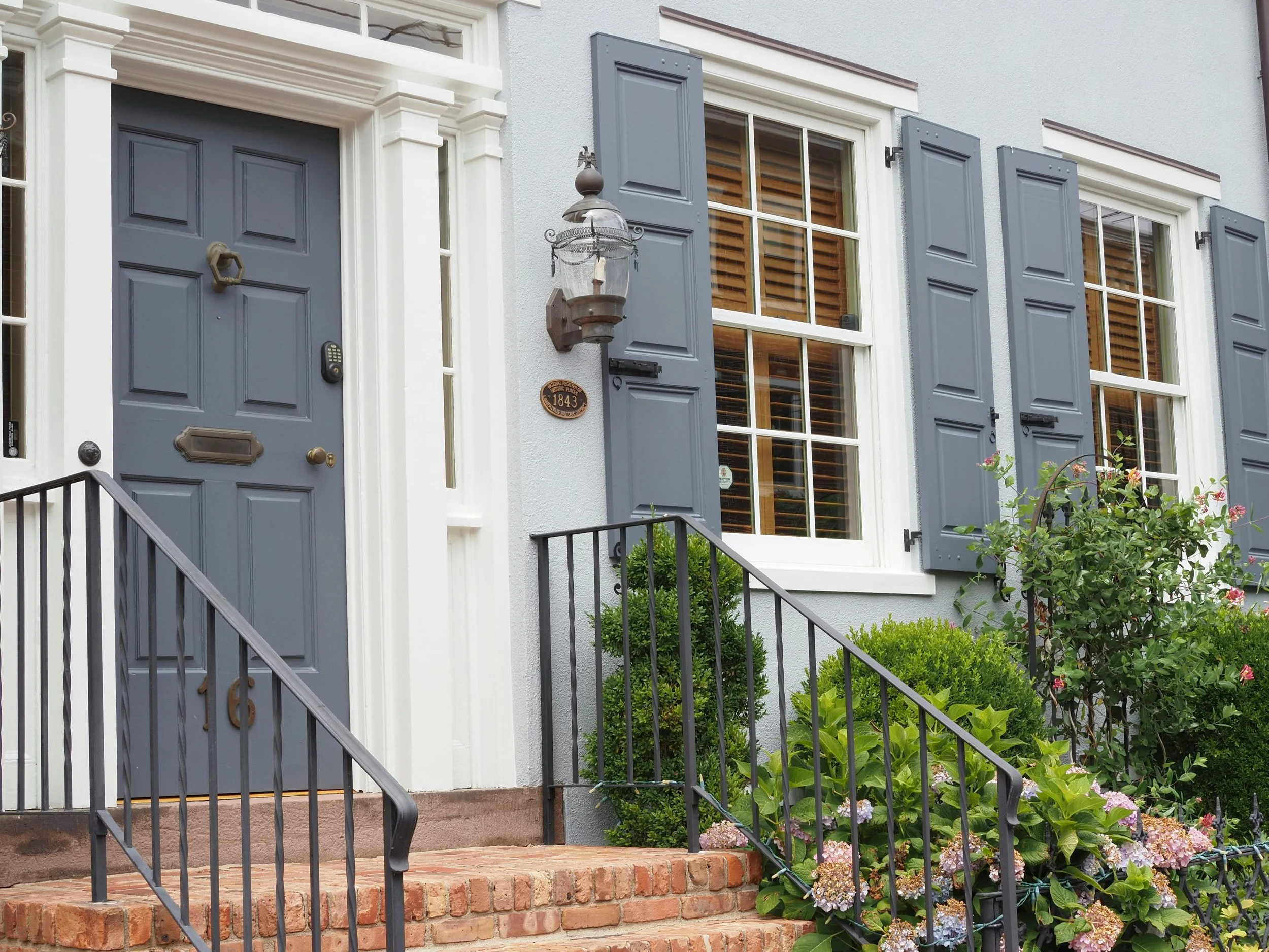 Front view of a house with a gray door, brick stairs, black railings, three windows with gray shutters, a vintage lantern, and lush green plants and flowers outside.