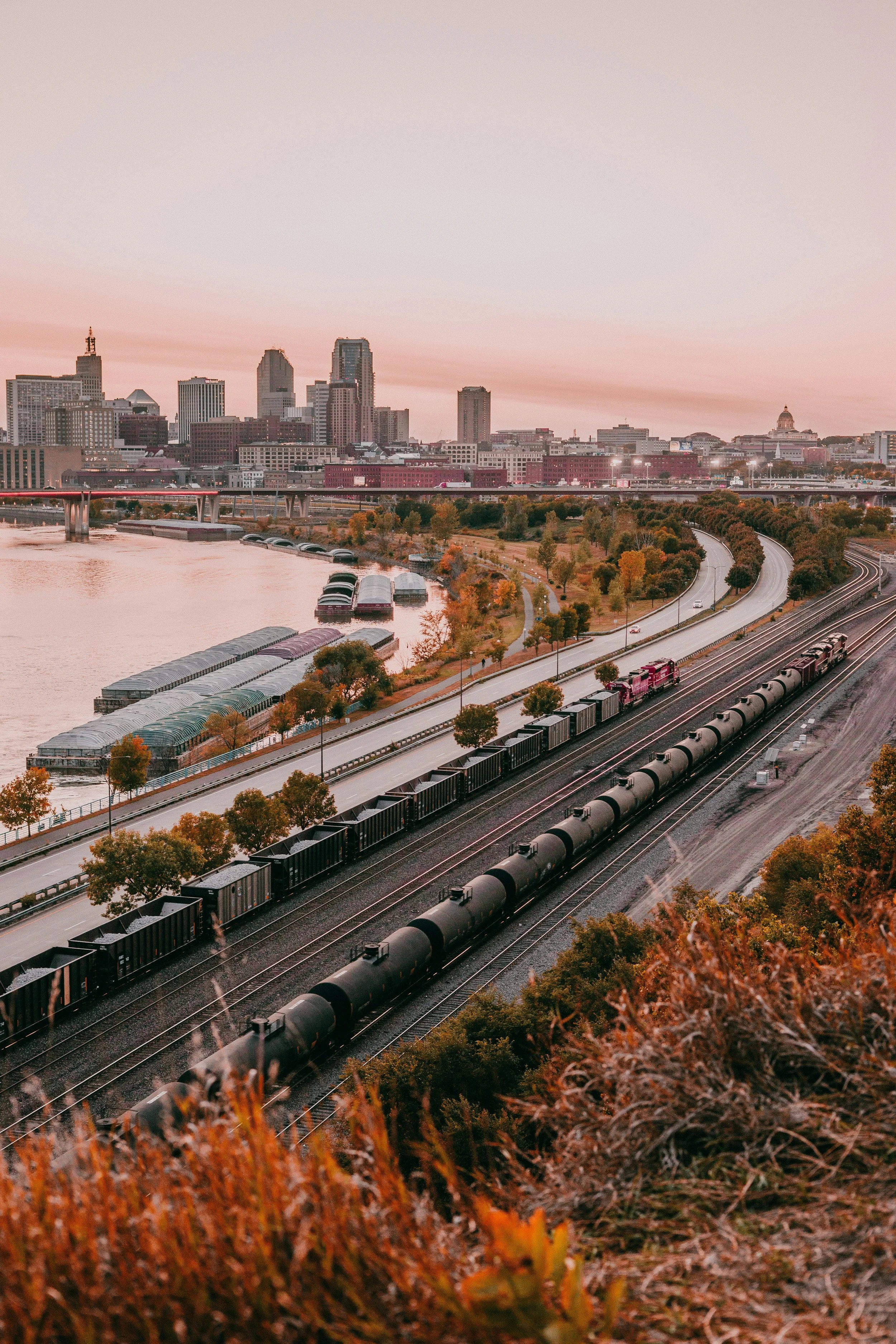 View of a train with black freight cars traveling along a railway track near a river in an urban cityscape during sunset, with tall buildings in the background and trees along the road.