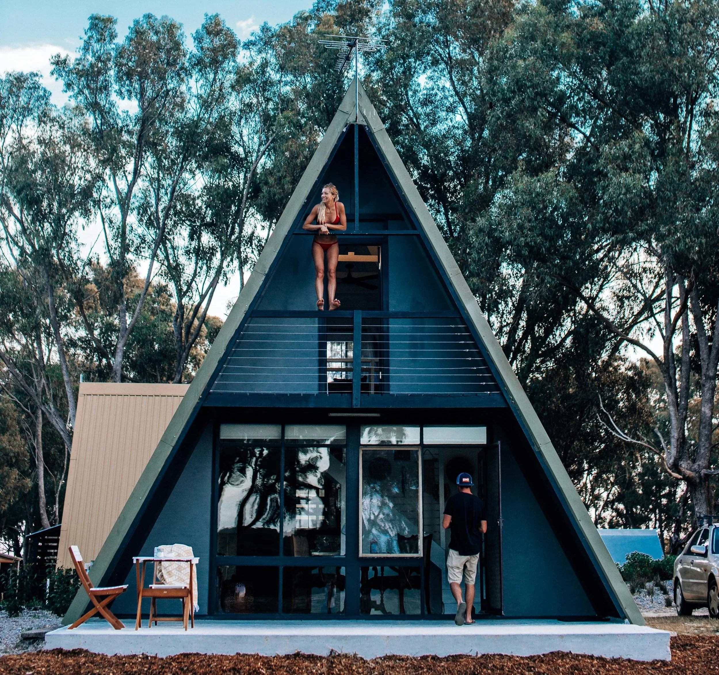 An A-frame house with a woman standing on the top balcony and a man standing at the entrance, with trees in the background and outdoor furniture in front.