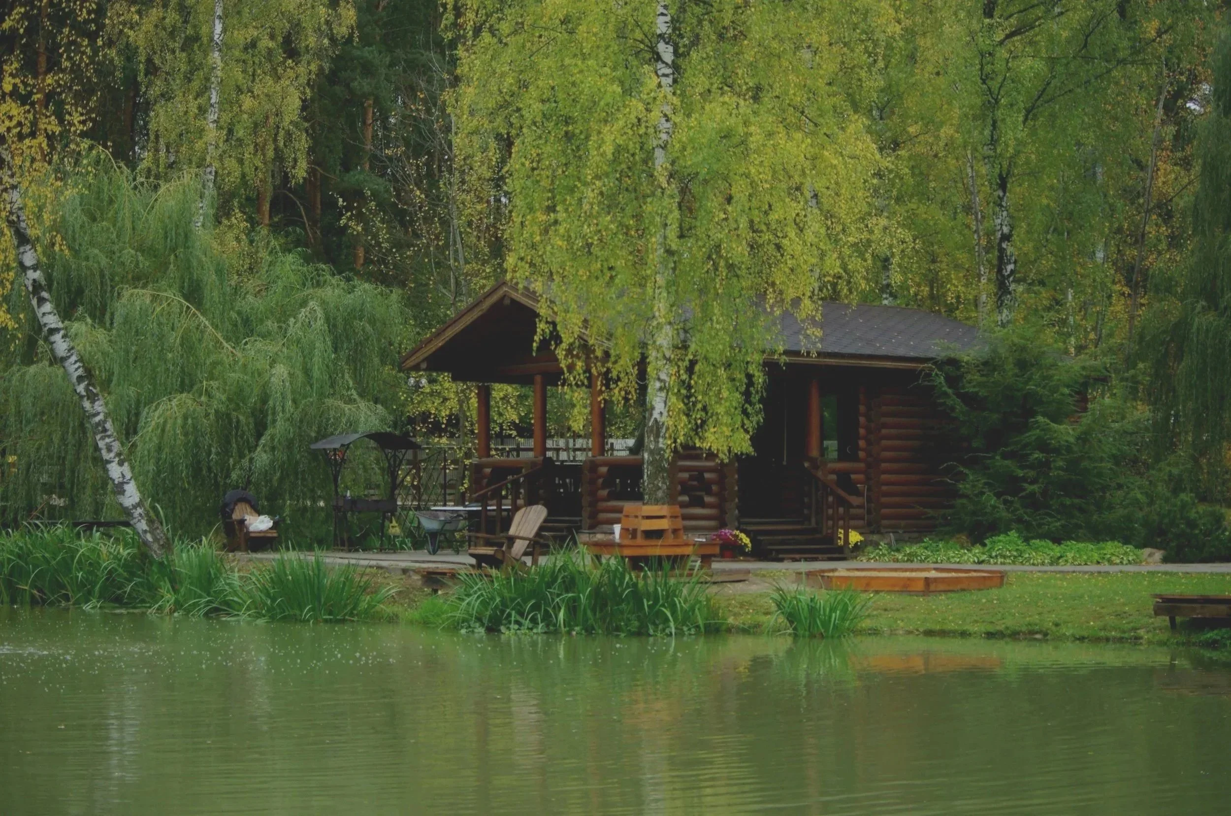 A wooden cabin by a pond, surrounded by trees with green leaves.