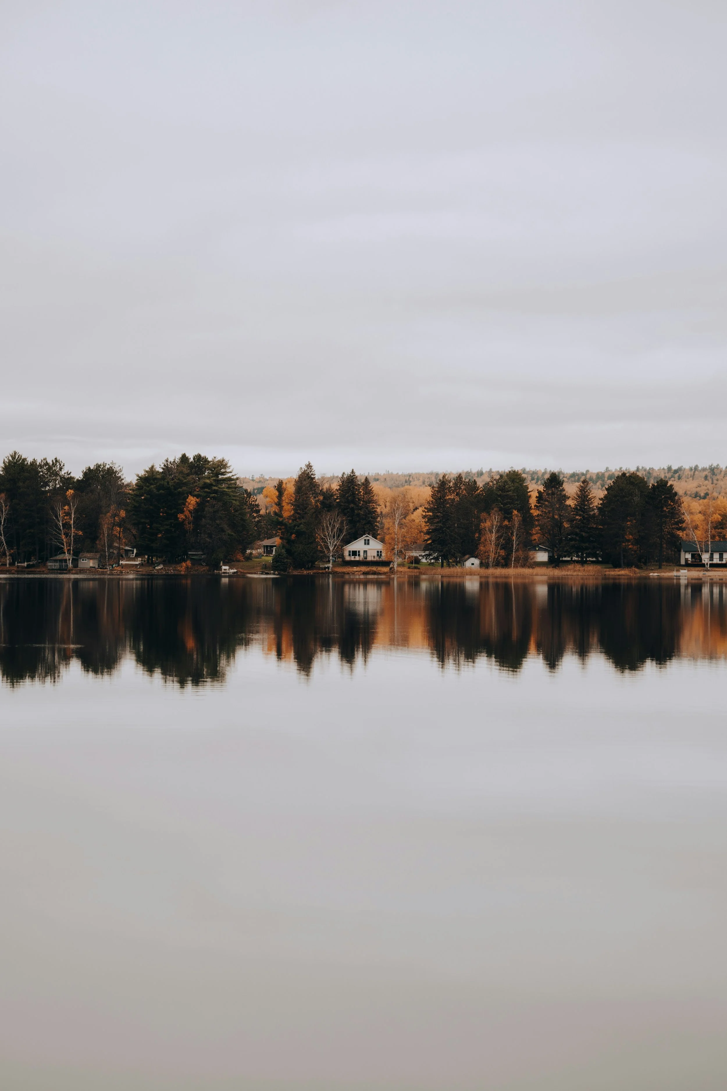 A calm lake reflecting a row of houses and trees along the shoreline, with an overcast sky.