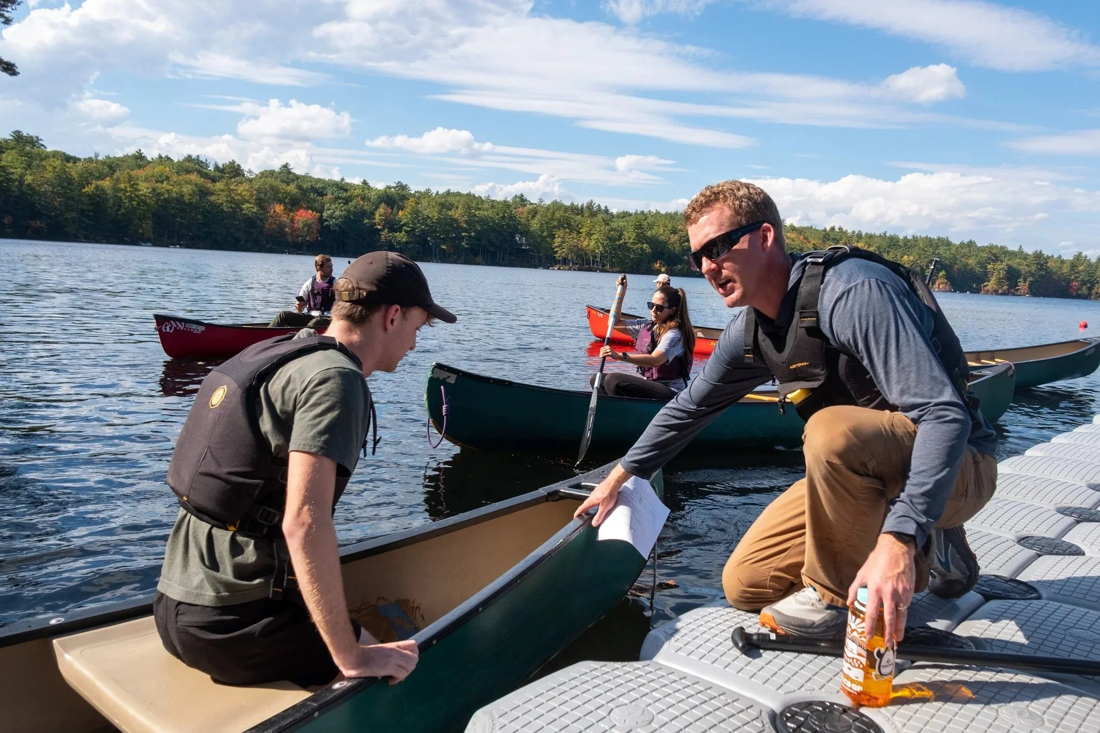 John Henkelman helping people preparing for canoeing on a lake with a dock, with a backdrop of trees and a sky with white clouds.