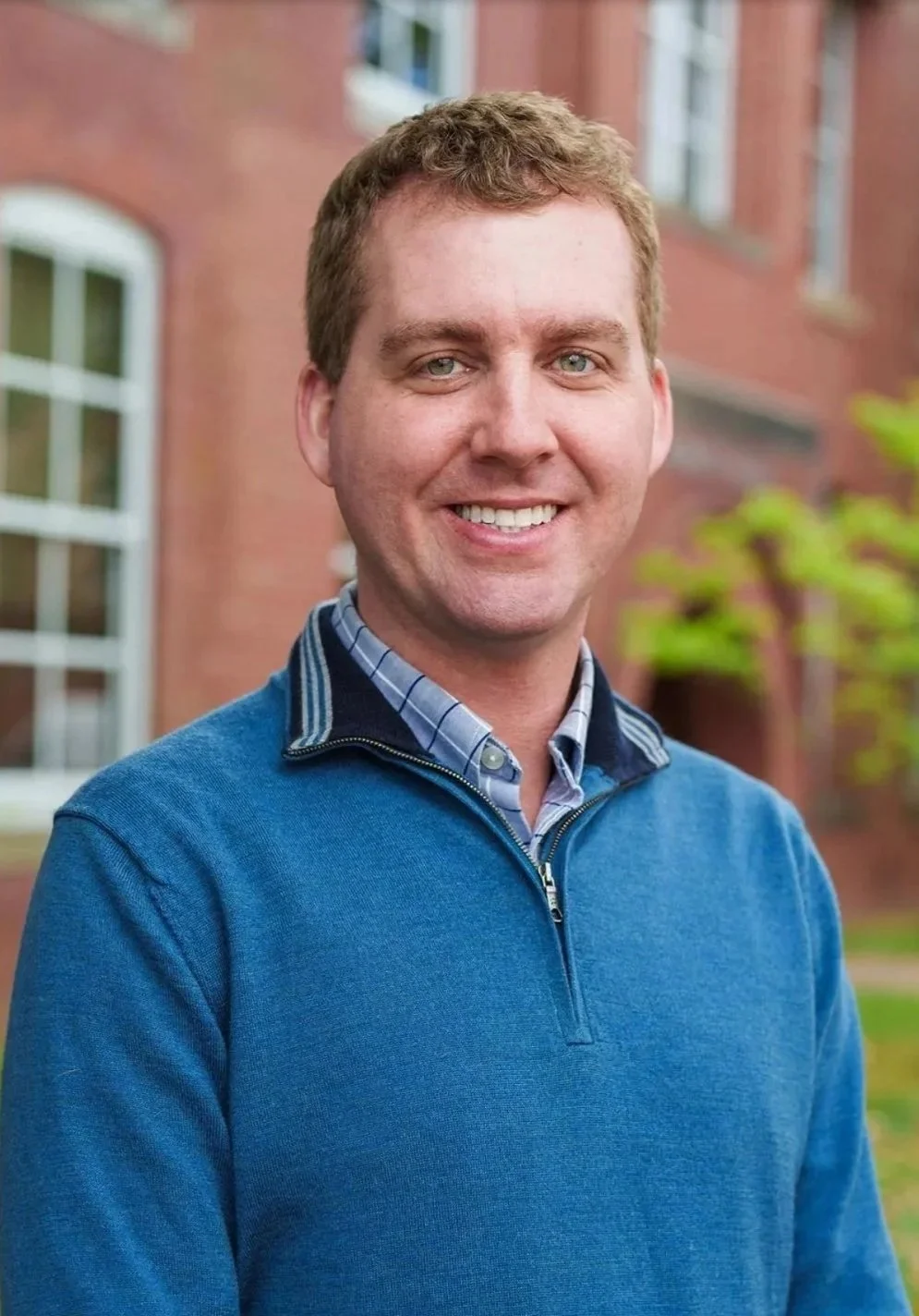 Portrait of a John Henkelman, a smiling young man with blonde hair and blue eyes, wearing a blue sweater and checkered shirt, standing outdoors in front of a brick building with windows and some greenery.