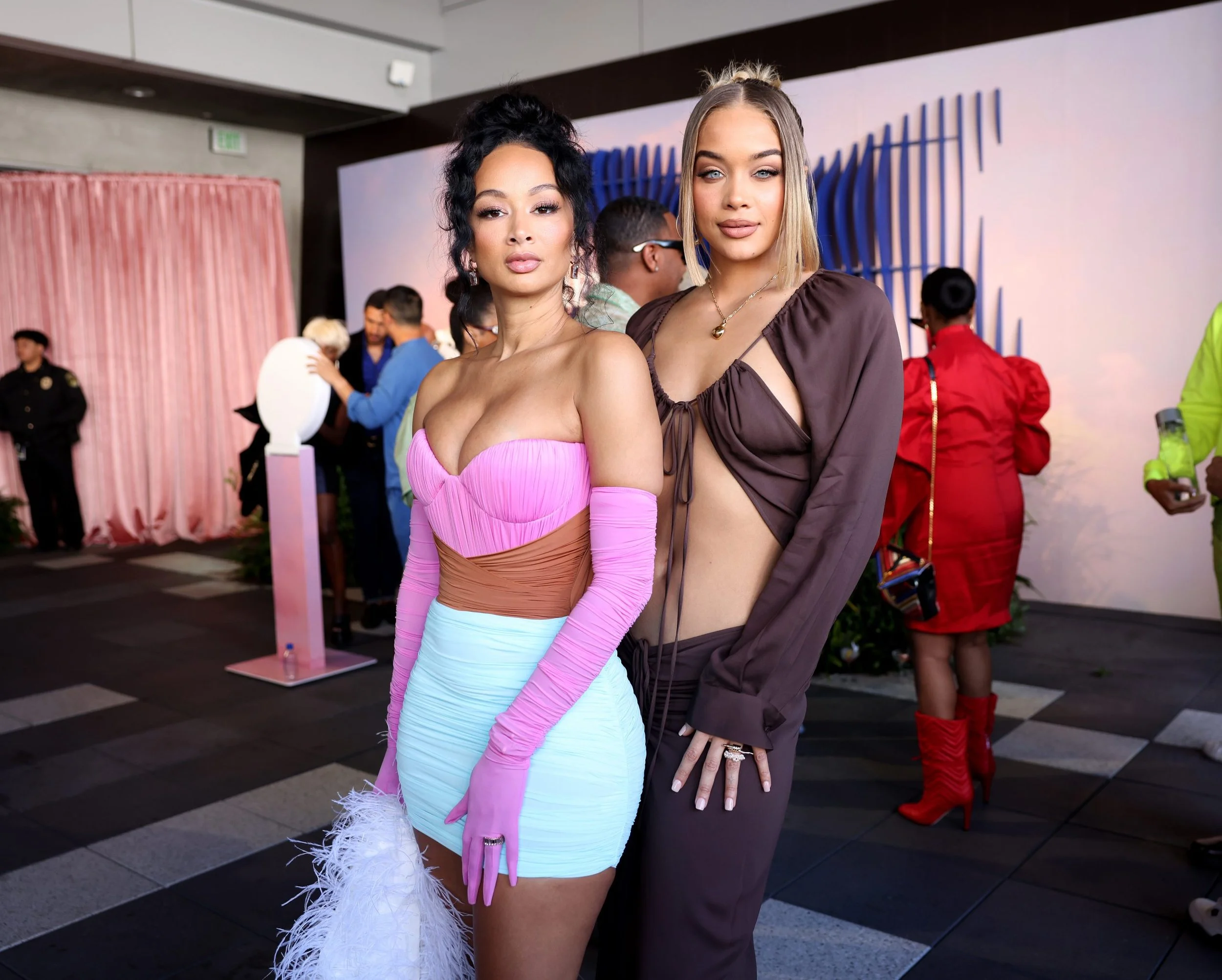 Two women posing at a social event, one wearing a colorful dress with long pink gloves and feathered accessory, the other in a brown cut-out dress with a necklace, with people and a large decorative backdrop in the background.