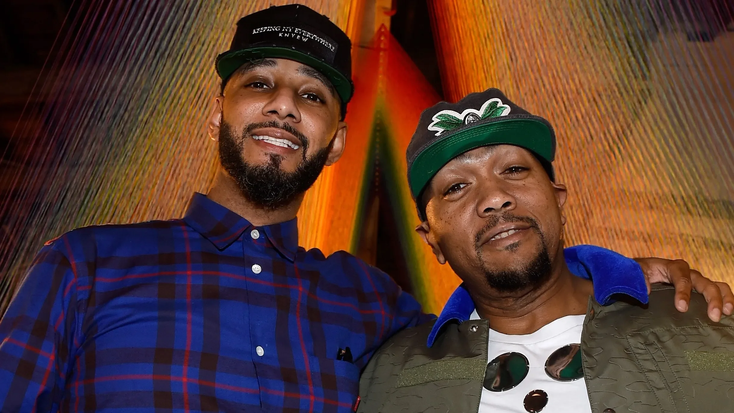 Two men standing closely together, smiling, with colorful abstract art background. The man on the left has a beard, wears a blue plaid shirt and a black cap. The man on the right has a goatee, wears a gray cap with green brim and a jacket over a whit