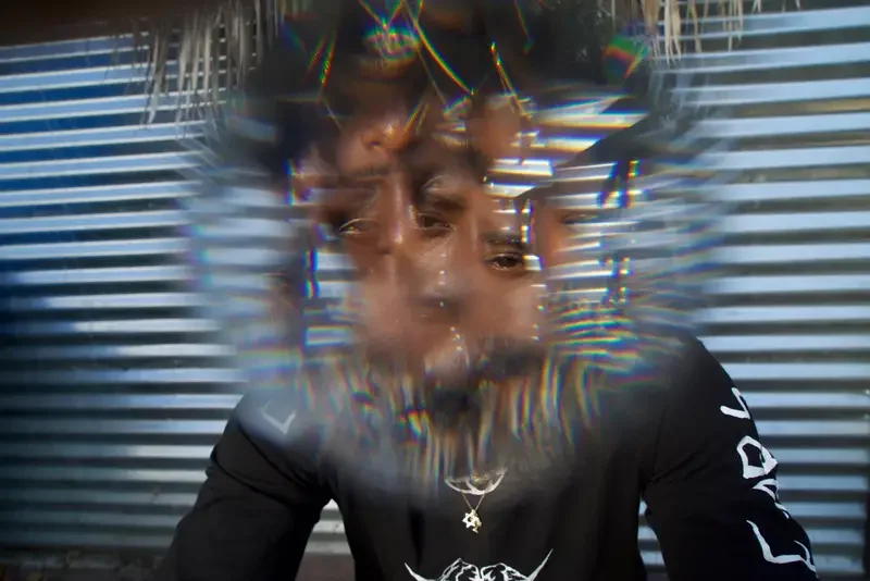 A person is seen through a distorted, kaleidoscope-like lens, creating multiple overlapping images of their face, with metal blinds in the background.