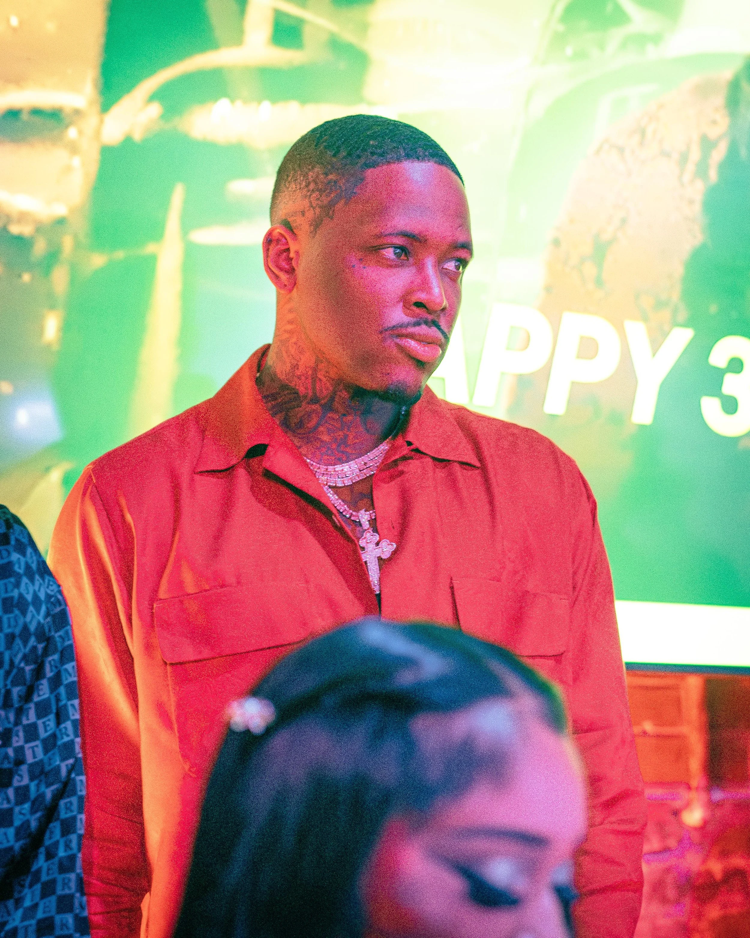 A young man with tattoos on his neck wearing a red shirt and layered necklaces, standing in front of a colorful background with partial text 'HAPPY 3' visible, at a vibrant social event.