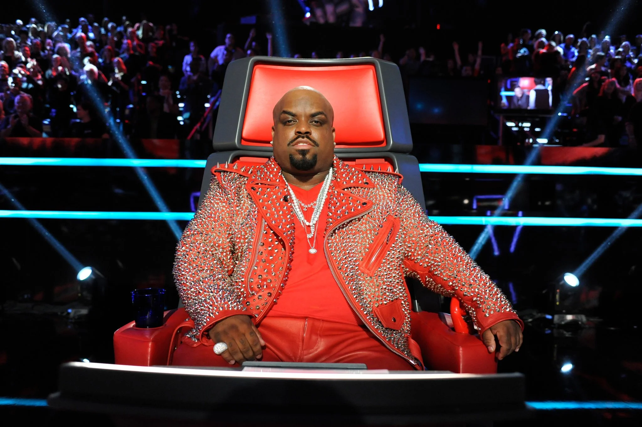 A man sitting on a large red chair on a stage, wearing a red studded jacket and jewelry, with an audience in the background.
