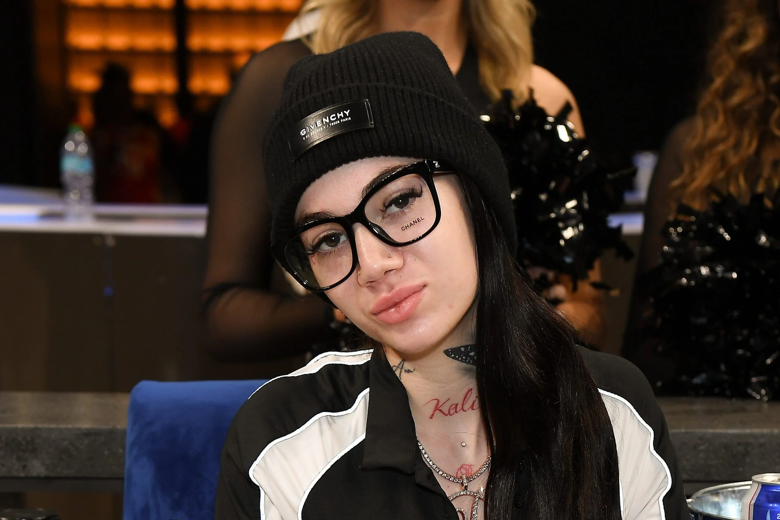 Young woman with long black hair wearing black glasses, a black beanie with a Givenchy logo, and a black and white jacket, sitting at a table in a dimly lit indoor setting.