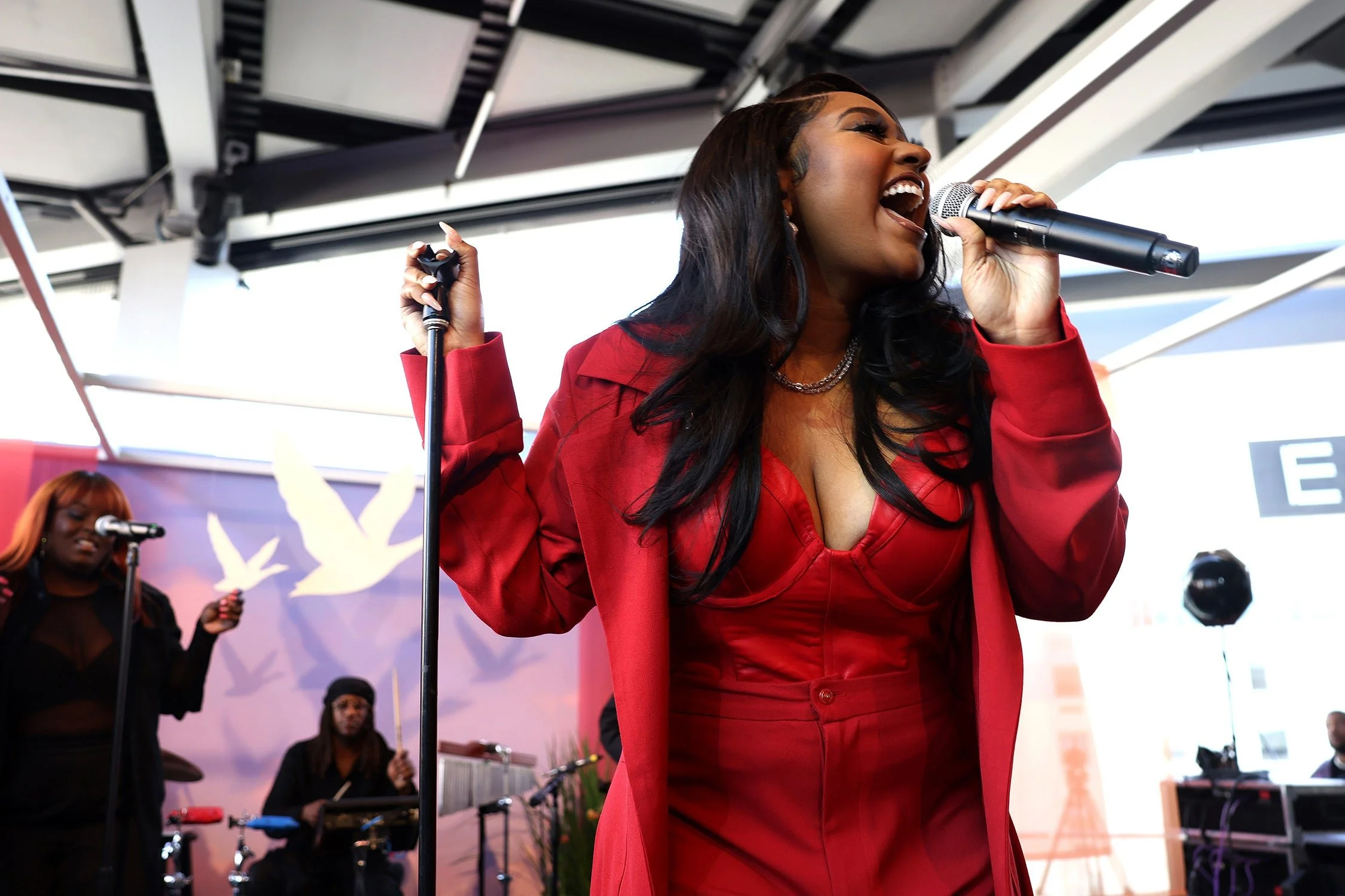 A woman singing passionately into a microphone on stage, dressed in a red outfit with a matching blazer, with other musicians playing in the background.