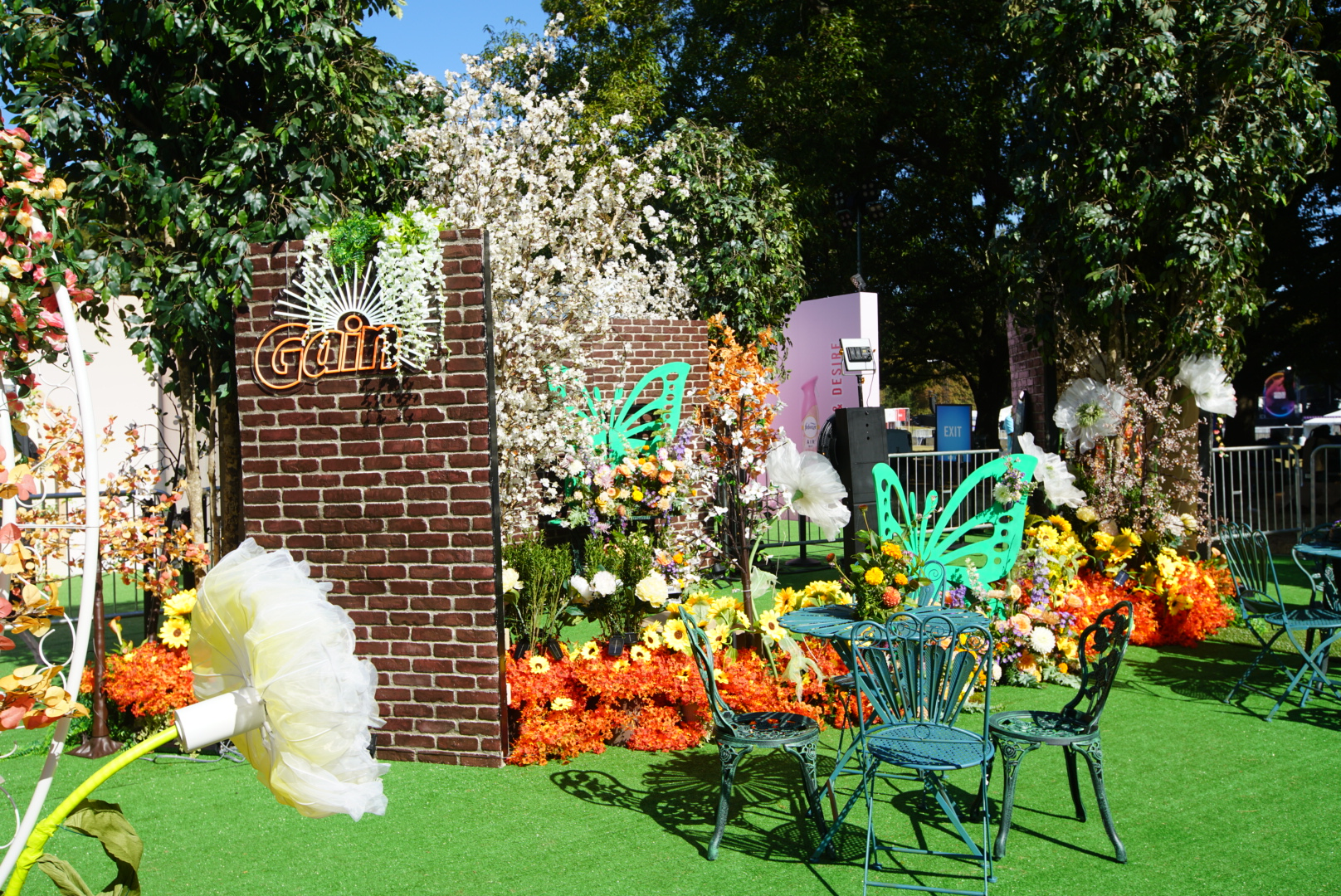 Outdoor decorative garden area with artificial grass, colorful flowers, metal chairs, a brick wall with a neon "GIRL" sign, and trees in the background