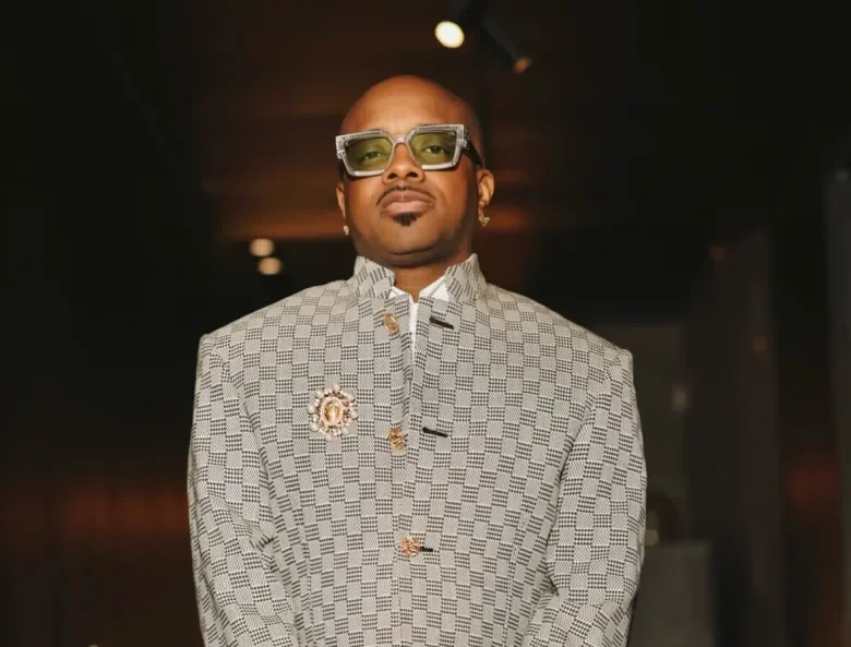 A man wearing large, stylish glasses and a patterned blazer with gold accents, posing in a dimly lit setting.