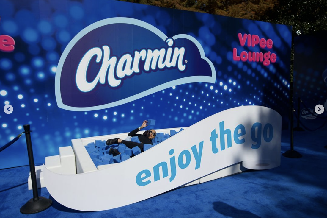 Person lying on a foam pit inside a Charmin-themed lounge with blue and white decorations, advertising 'enjoy the go' and 'VIPEe Lounge'.